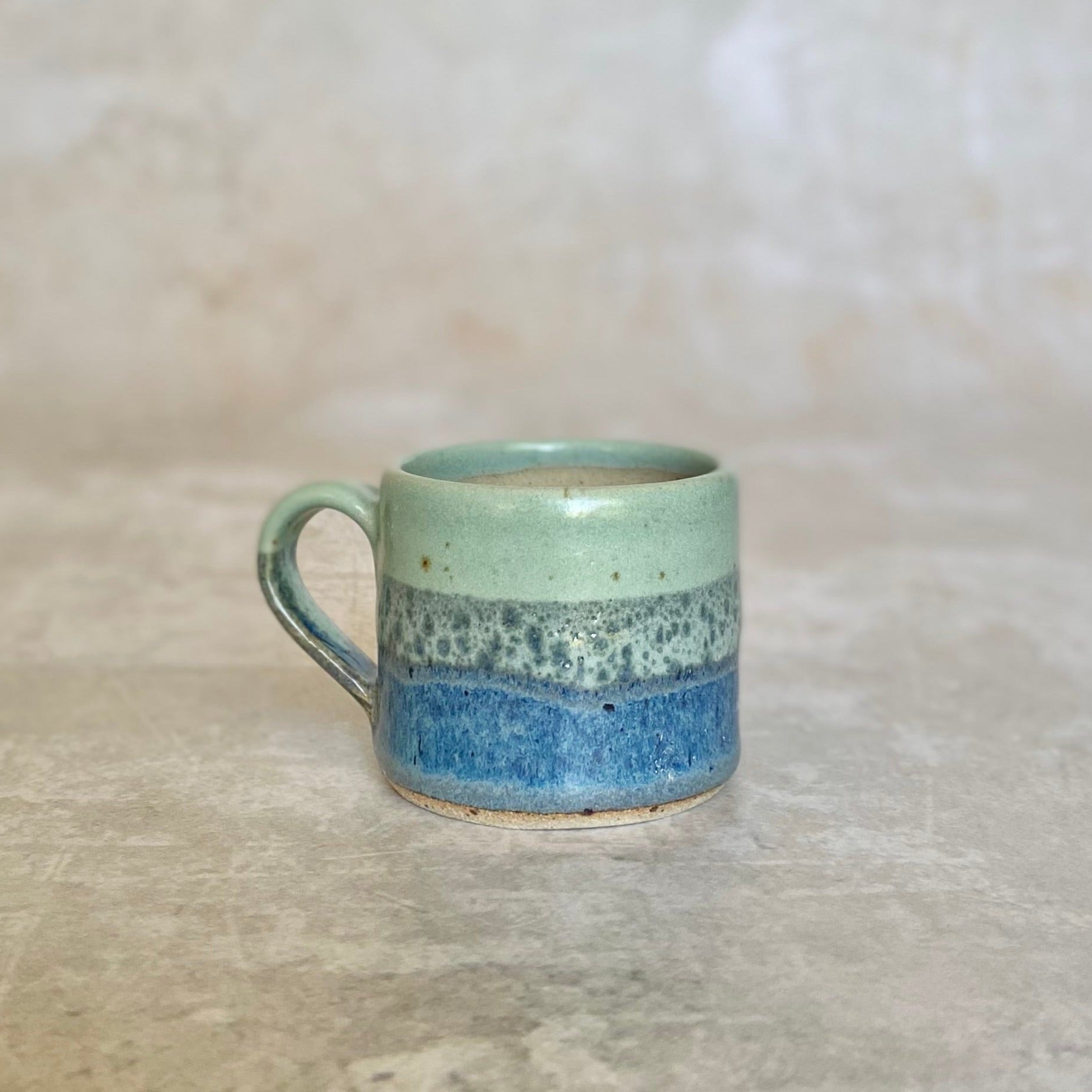 Small ceramic mug with a light green, speckled light teal, and dark teal glaze, in horizontal bands.