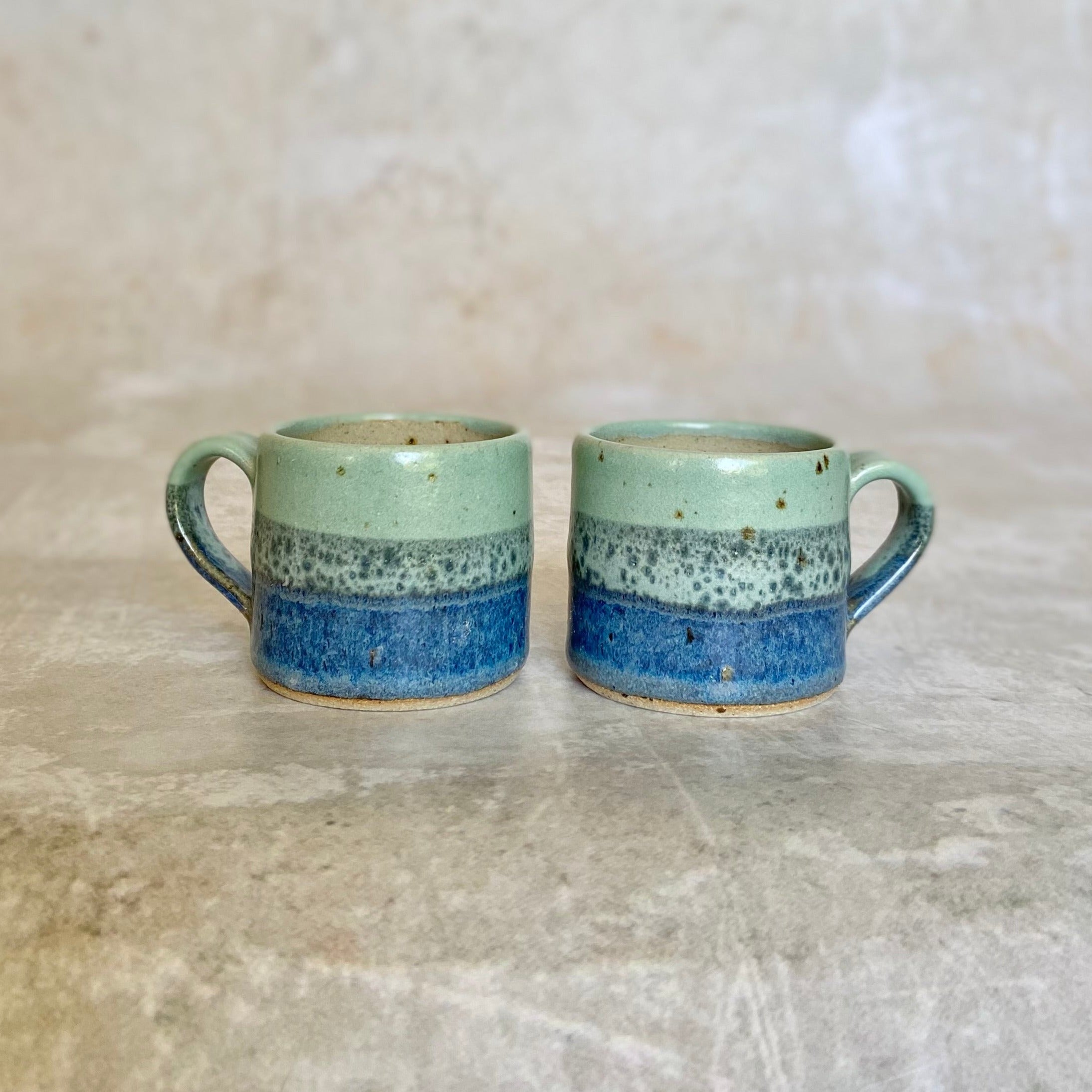 Pair of small ceramic mugs with light green, speckled gray, and dark blue horizontal bands.