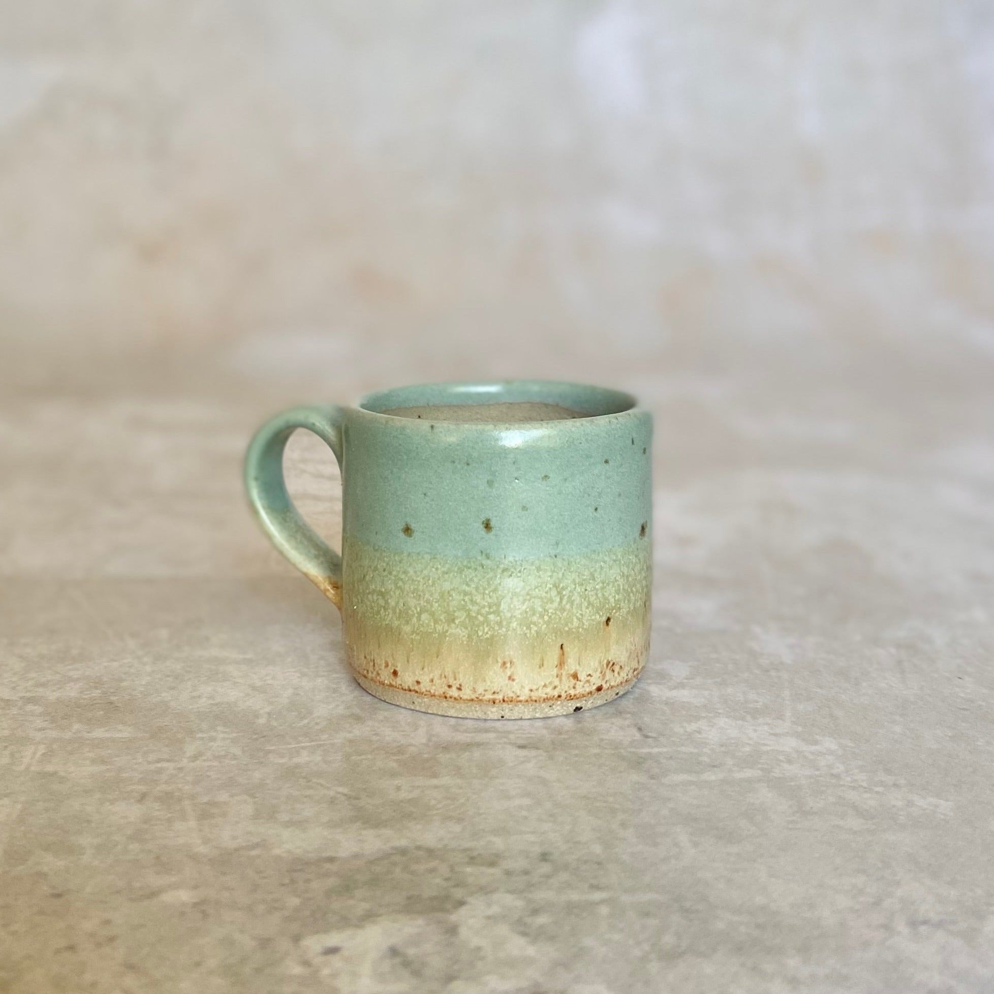 Small ceramic mug with a light teal and beige speckled glaze.  The mug has a rounded handle.