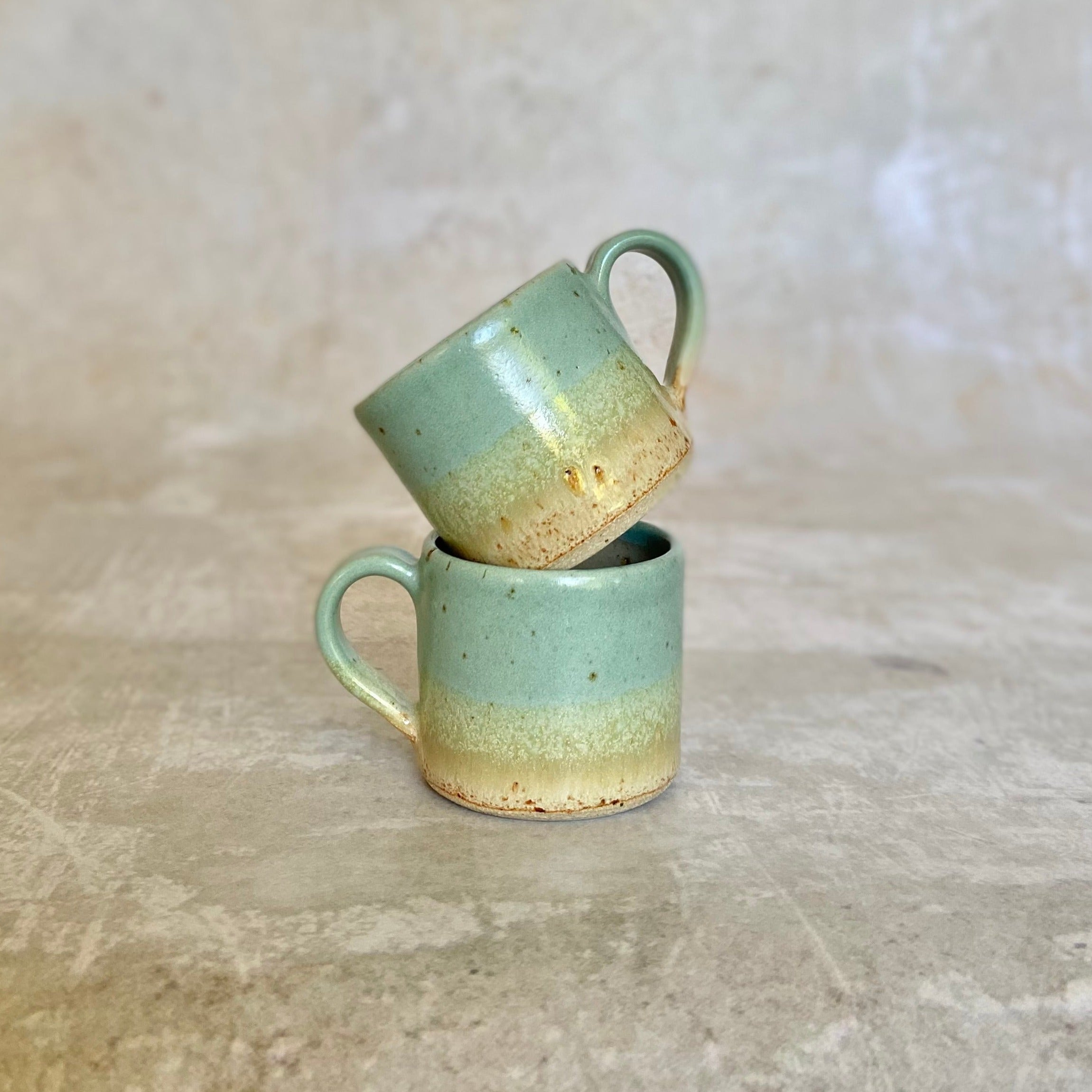 Two stacked ceramic mugs; light teal glaze with a speckled, beige bottom band.