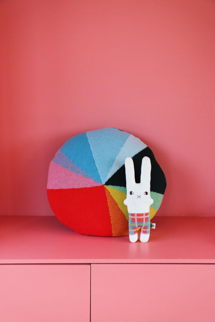 A colorful round cushion by Donna Wilson with a patchwork design, next to a white bunny plush in plaid pants.