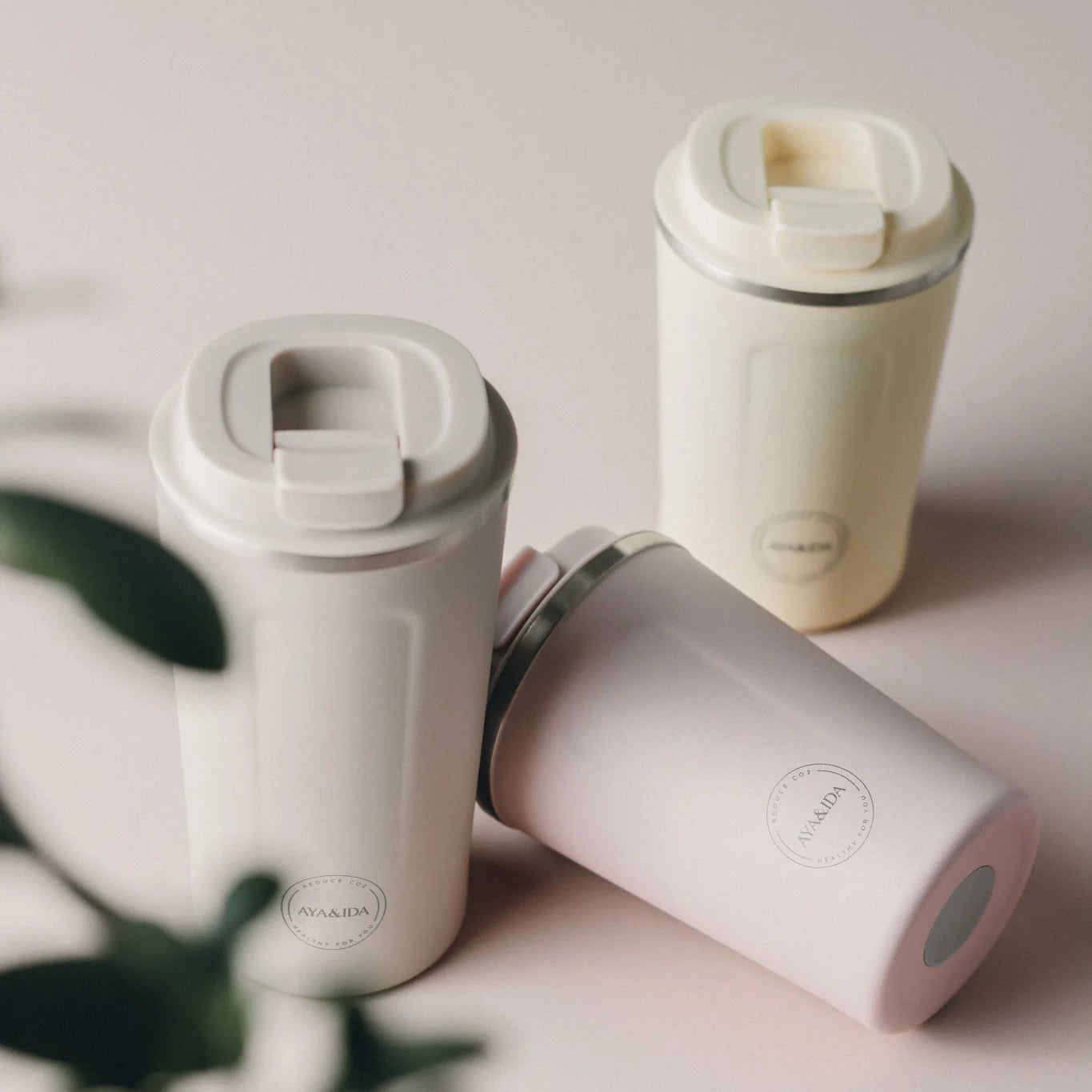 Three reusable coffee cups in beige and pink; each has a press-fit lid and a subtle brand logo.
