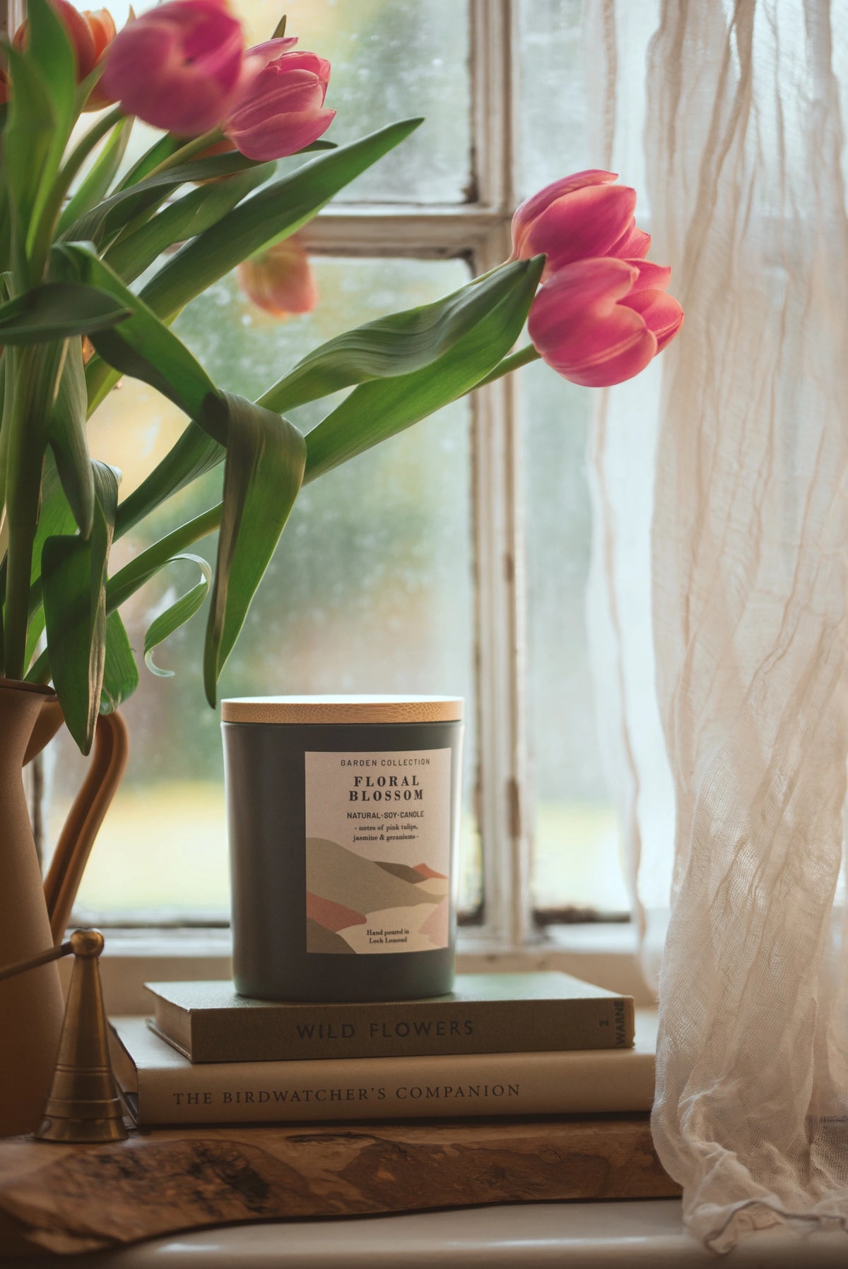 A jar candle labeled 'Floral Blossom' sits on stacked books on a windowsill, with pink tulips in a vase and sheer white curtains beside it.
