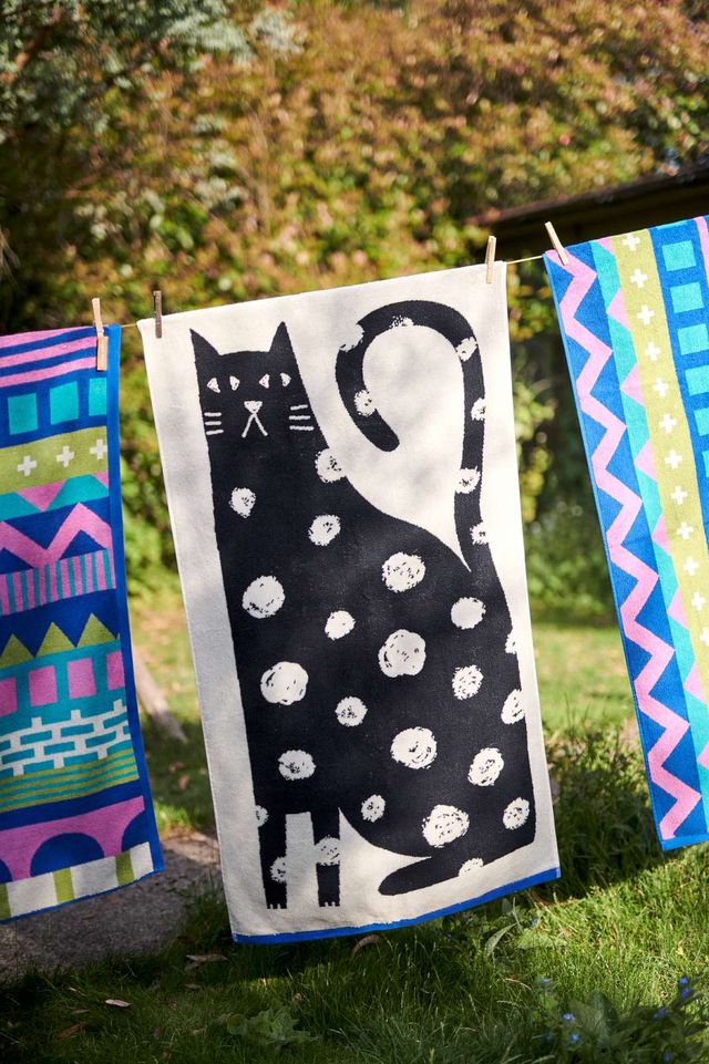 A white kitchen towel with a black cat silhouette pattern hanging outdoors on a clothesline between wooden clips, flanked by two colorful patterned towels against a blurred green garden background.