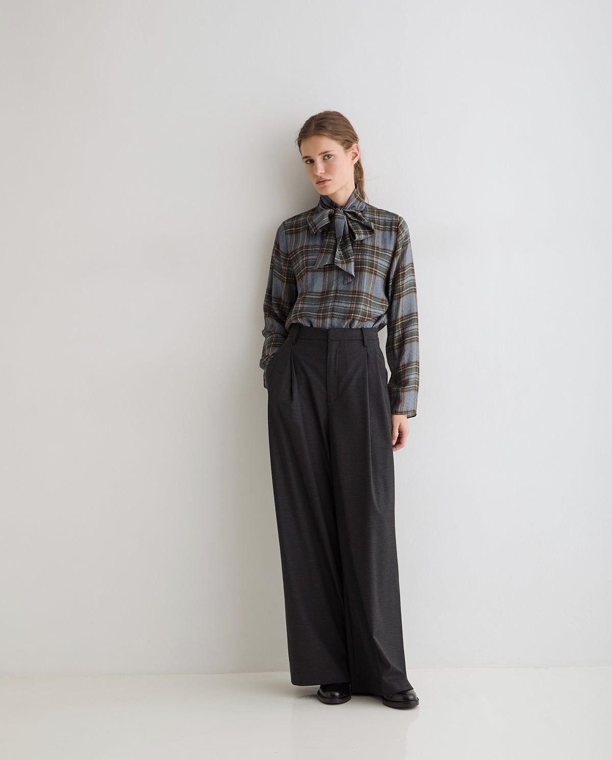 A model stands against a plain light gray studio wall wearing a long-sleeve plaid button-up shirt with a large bow tie collar in muted blue, gray, and brown tones, paired with high-waisted black wide-leg trousers and black shoes.