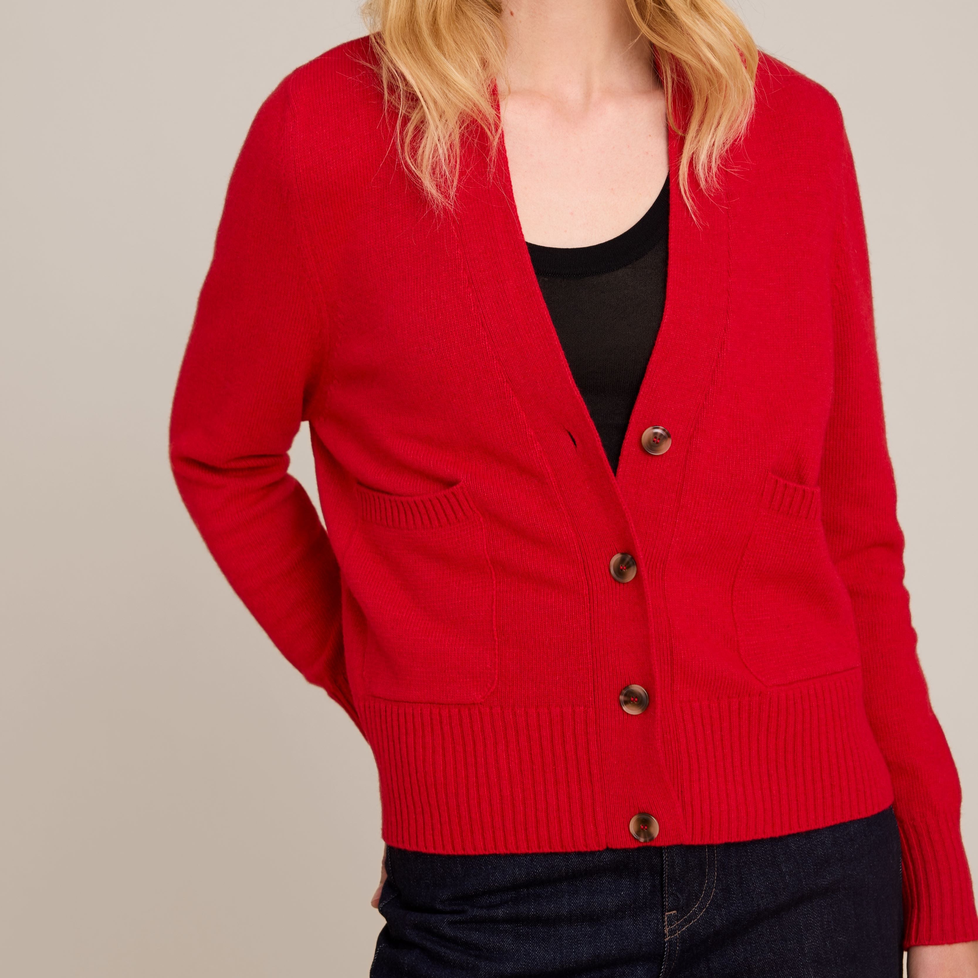 A woman wearing a bright red open-front cardigan with dark buttons, layered over a black top and dark jeans, photographed against a plain light gray studio background.