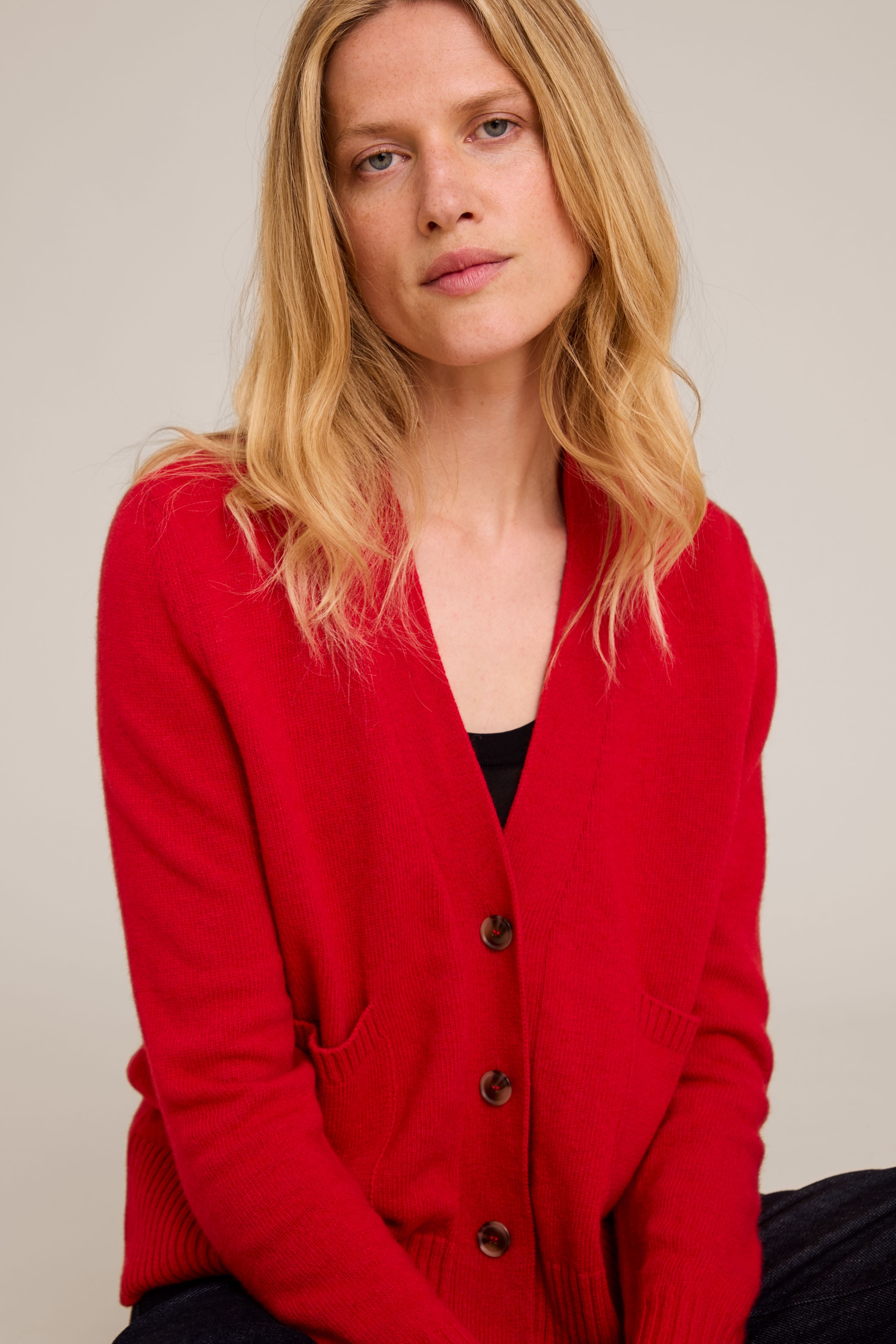 A blonde woman wearing a bright red open-front knit cardigan with dark buttons over a black top, photographed against a plain light beige studio background.