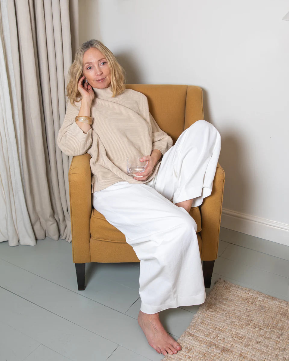 Woman sitting in a yellow armchair holding a glass, wearing a beige sweater and white pants.