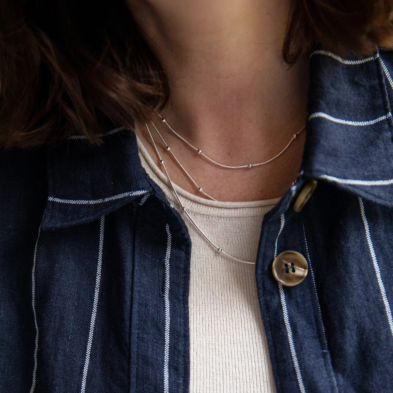 Person wearing a navy blue striped shirt over a beige sweater with a necklace.