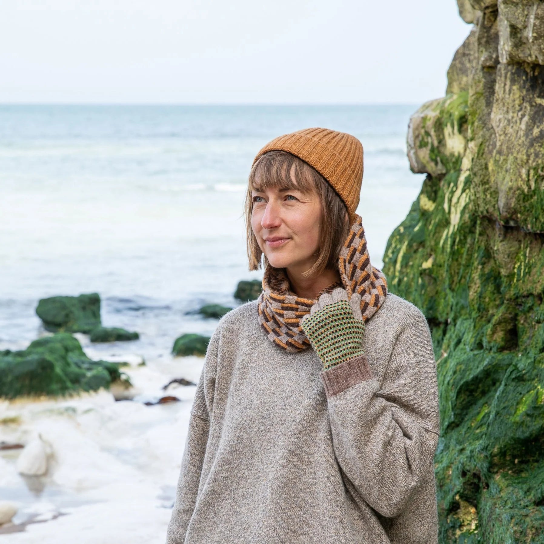 Woman wearing a light gray sweater, orange knit hat, patterned scarf, and fingerless gloves near the ocean.