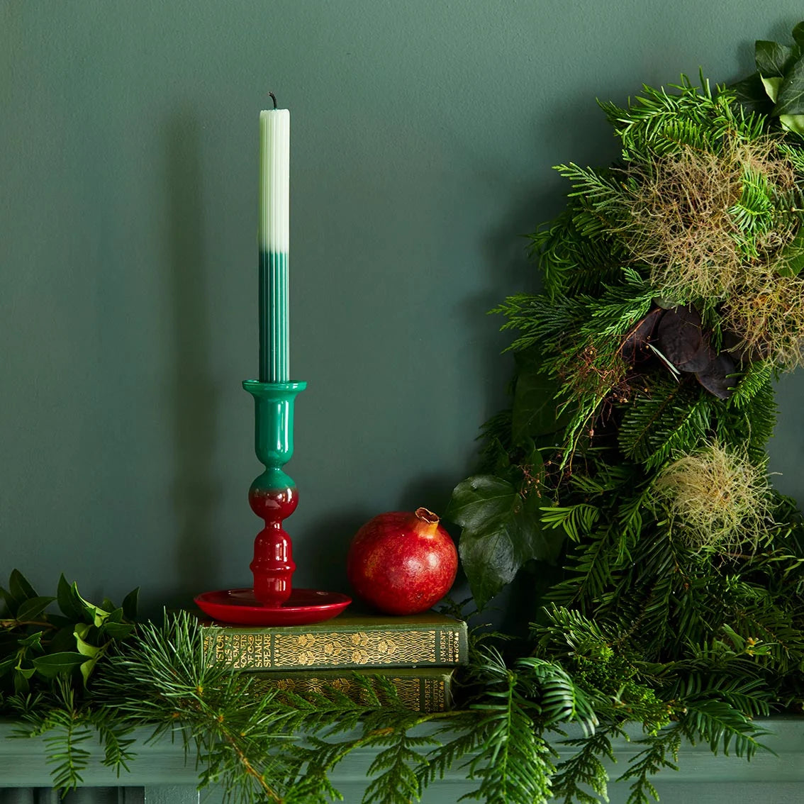 Decorative candlestick with a green and red design, pomegranate, and greenery against a dark green wall.
