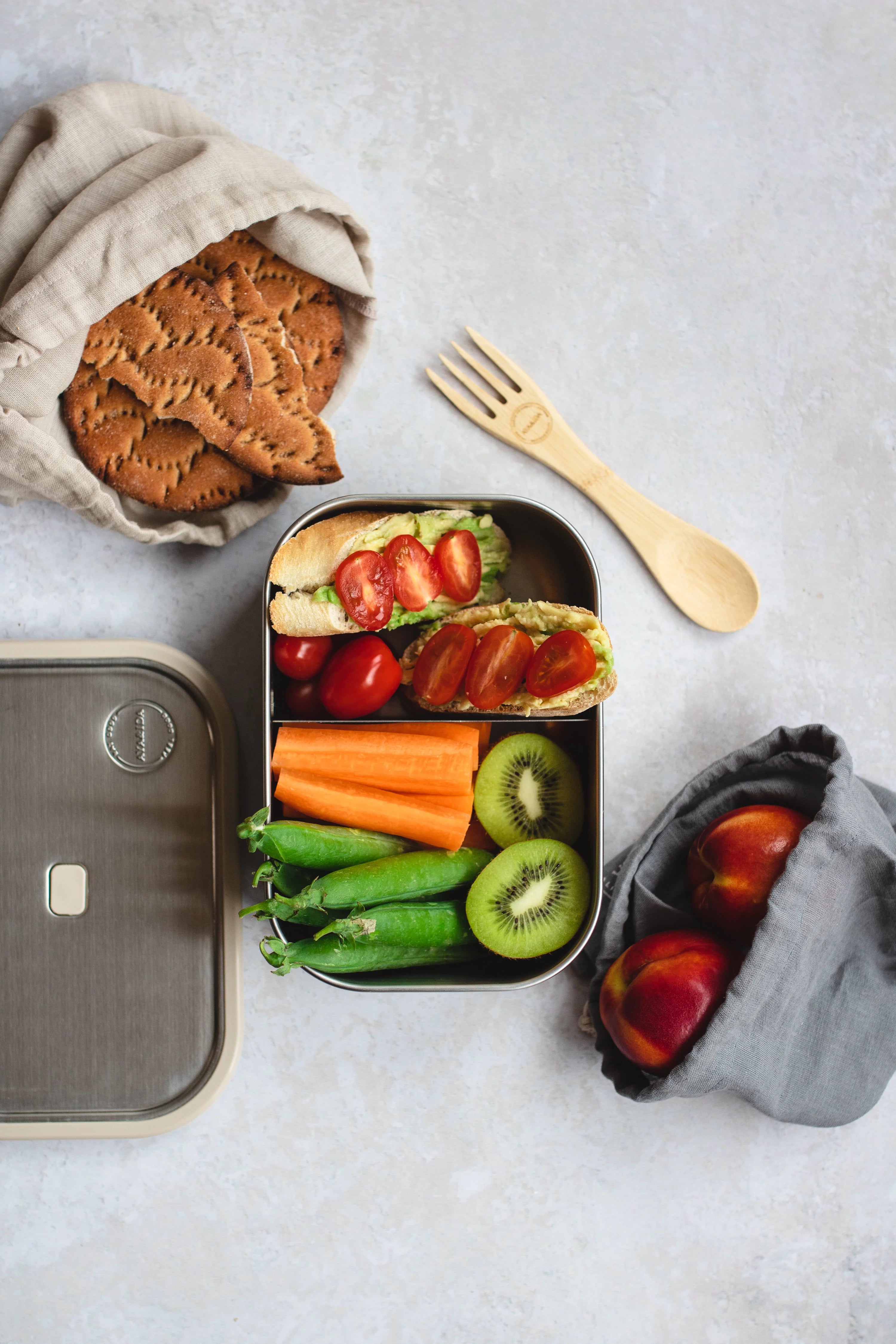 Stainless steel lunchbox with avocado toast, cherry tomatoes, carrots, peas, and kiwi.