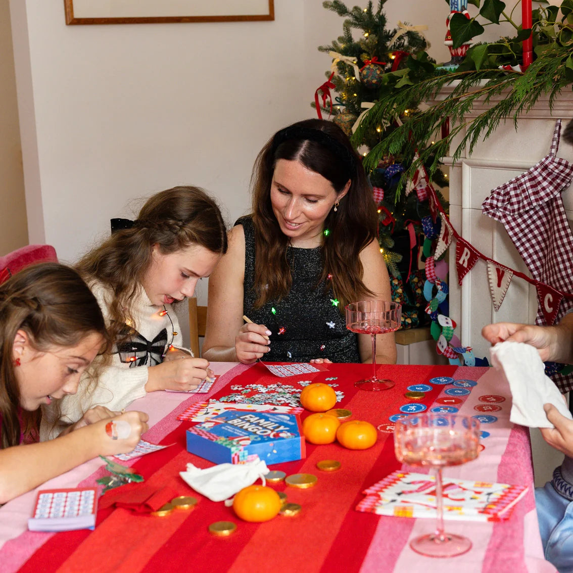 A woman and two girls are gathered around a table playing a card game during Christmas. The table is covered with a red and pink striped tablecloth, decorated with oranges, playing cards, and game pieces.