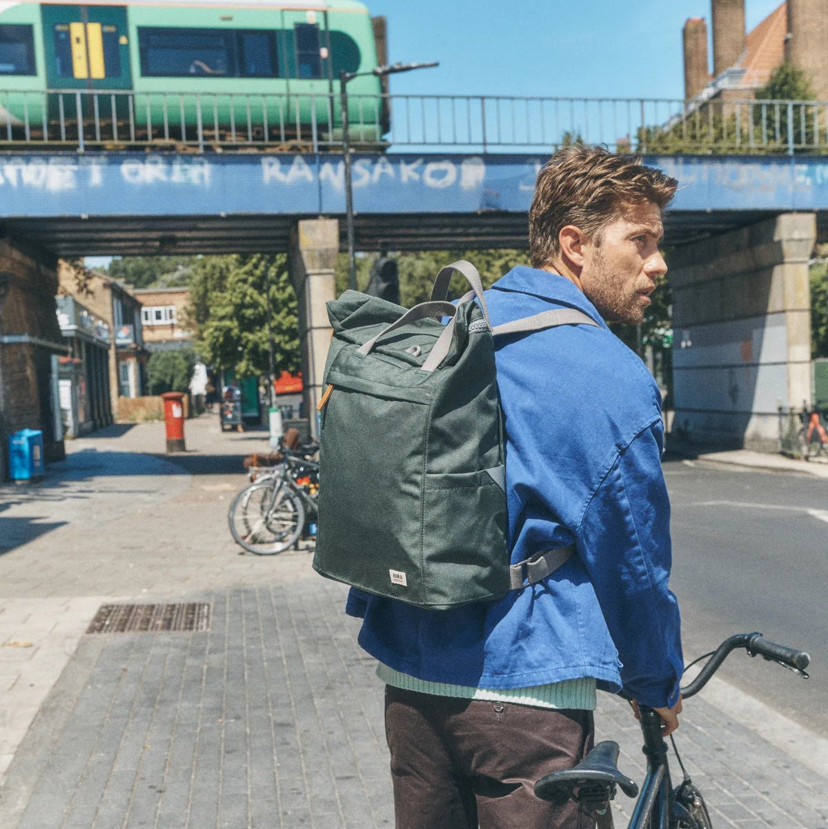 A man wearing a blue jacket and brown trousers stands on a city sidewalk next to a bicycle, carrying a large dark green backpack. An elevated train passes in the background under a bridge.