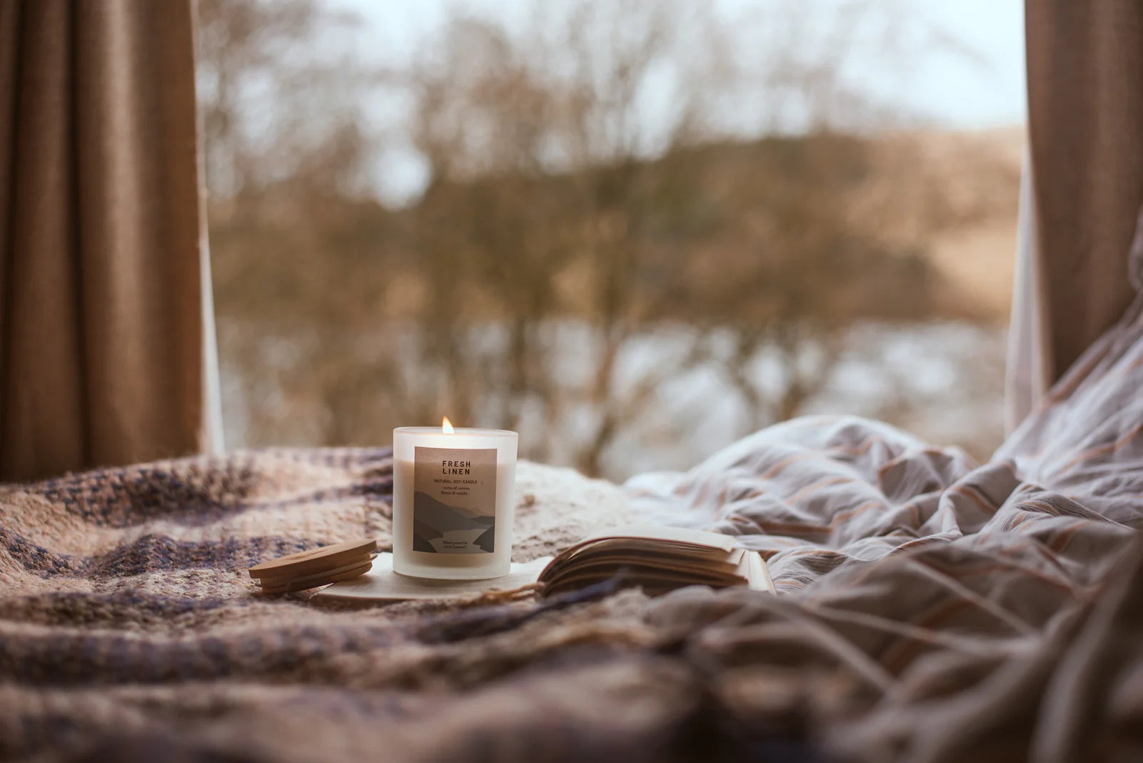A lit "Fresh Linen" natural soy candle in a frosted glass jar sits on a cozy blanket near an open book.