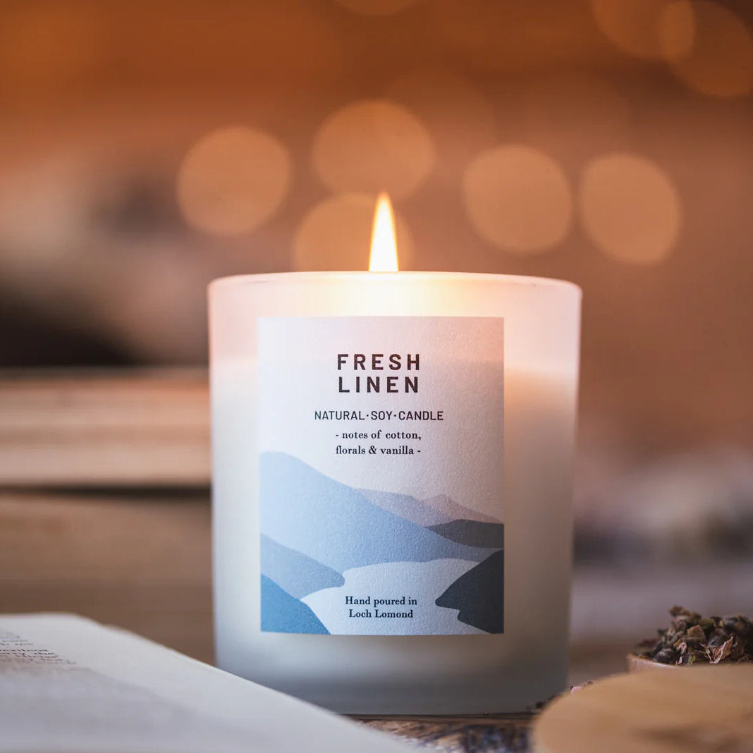 Lit frosted glass soy candle labeled "Fresh Linen," with notes of cotton, florals, and vanilla; hand-poured in Loch Lomond.