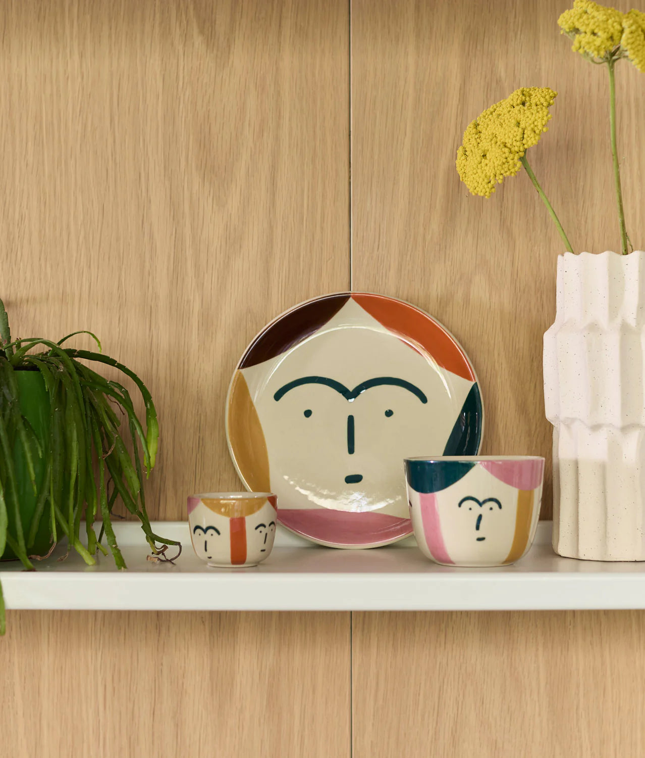 Ceramic face-themed plates and bowls on a wooden shelf with a plant and vase in the background.