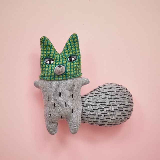 A handmade plush toy shaped like a fox, featuring a green and yellow patterned knit head with embroidered eyes and nose, a gray knit body with black stitched details, and a gray patterned tail, set against a solid pink background.