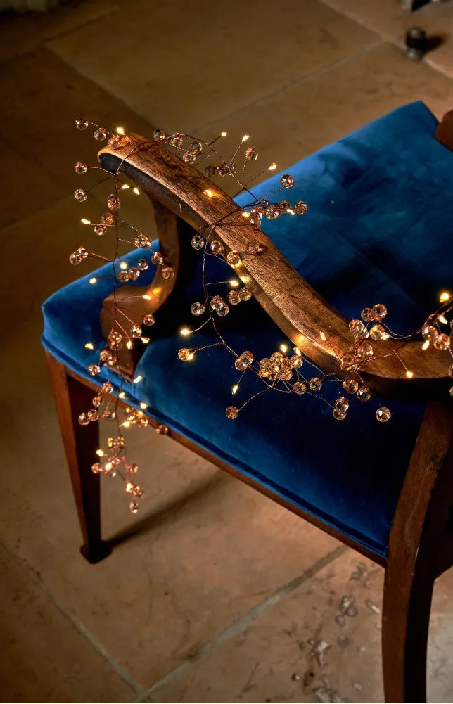 A blue velvet upholstered wooden chair in a warm indoor setting, decorated with a copper-toned branch wrapped in warm fairy lights and clear crystal-like beads.