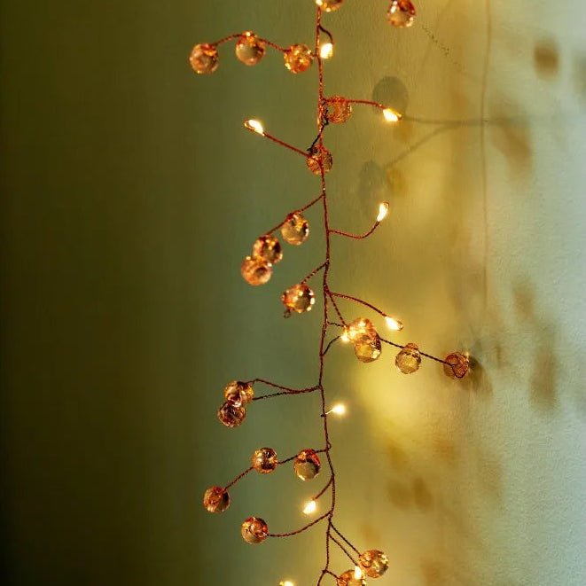 A decorative string of warm white fairy lights with clear faceted crystal-like beads on a thin brown wire, shown hanging against a softly lit wall.