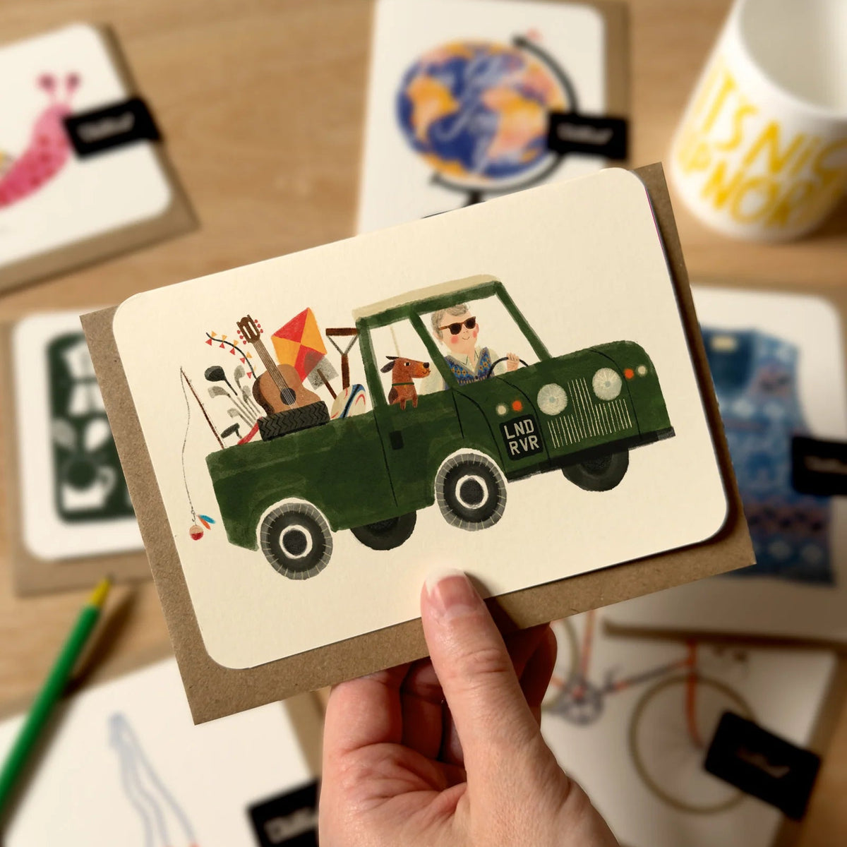 Hand holding a card with a green car illustration on a wooden surface with other cards and a mug.