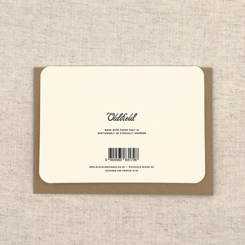 The back of a square kraft paper card or packaging insert on a textured beige linen background. The card has a cream-colored center with black text reading 'Oldfield' and a barcode.