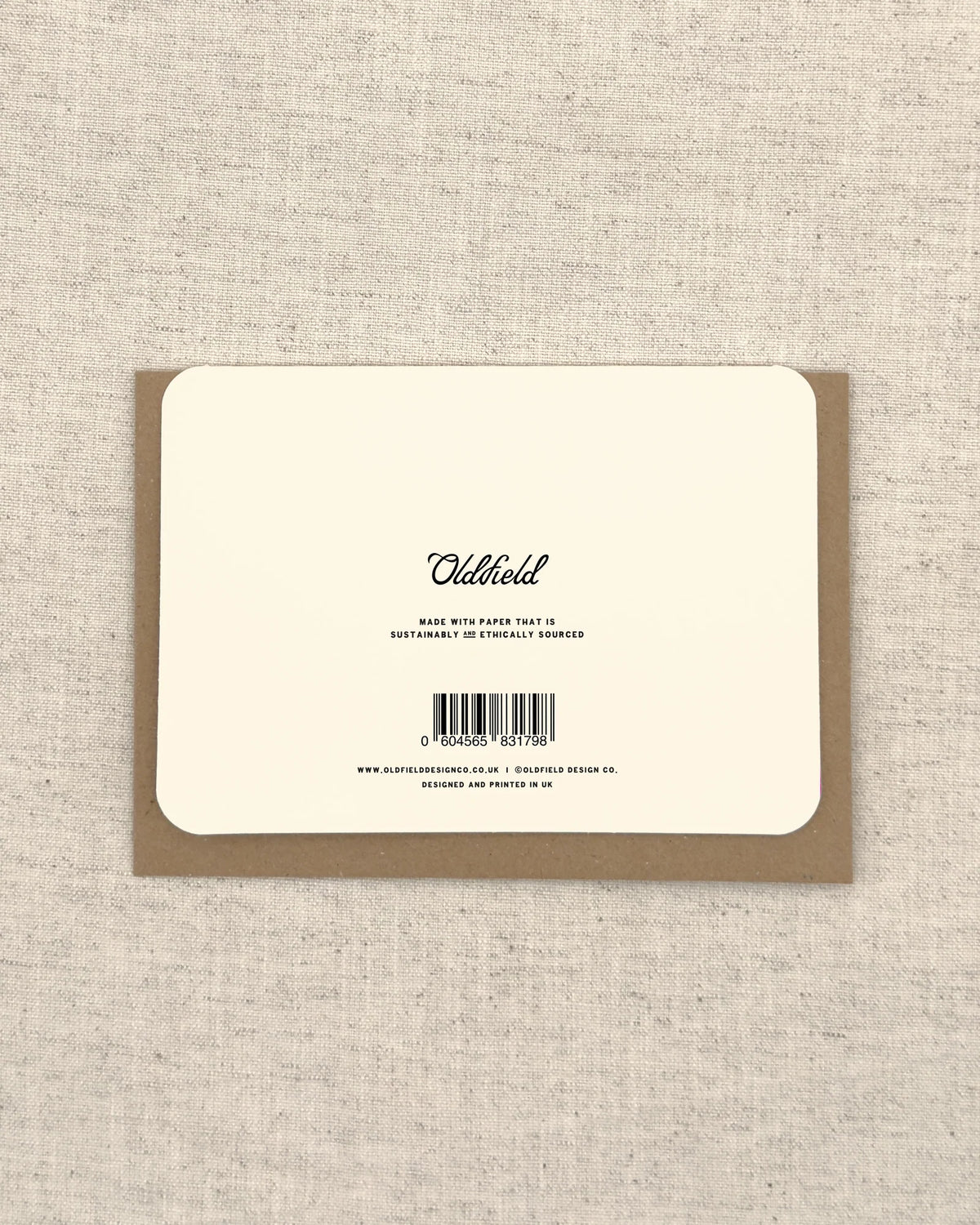 The back of a square cream-colored product card or packaging insert with black text reading 'Oldfield' and a barcode, placed on top of a brown kraft envelope against a textured beige linen background.