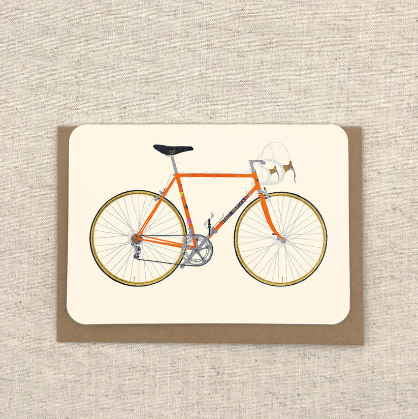 A square art print featuring an orange road bicycle with a small white basket, displayed on a brown kraft envelope against a textured beige linen background.