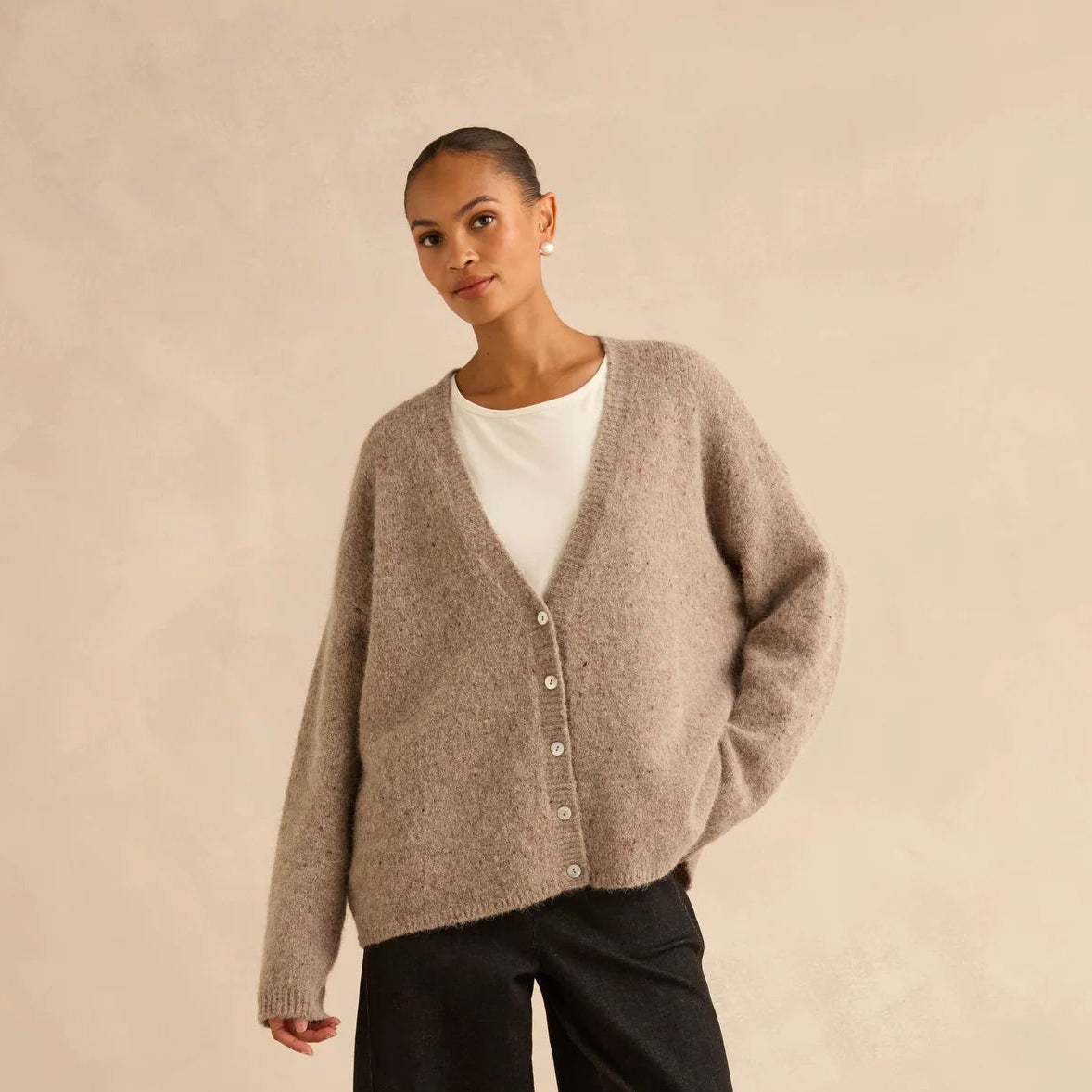A model stands against a warm beige studio backdrop wearing a taupe knit button-front cardigan over a white top, black wide-leg pants, and black boots.