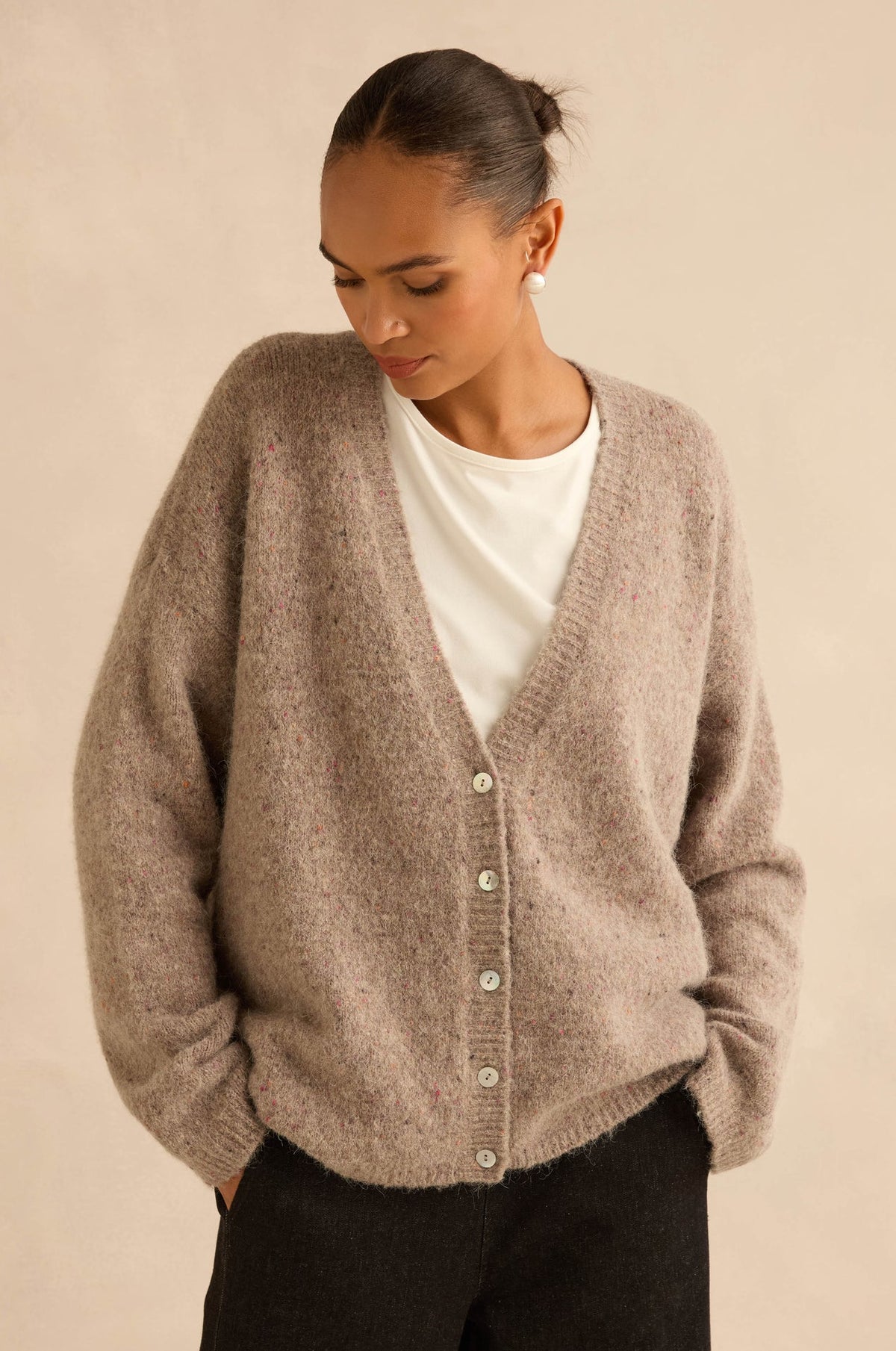 A woman wearing a fuzzy, oversized taupe knit cardigan over a white t-shirt and black pants, standing against a solid beige background.