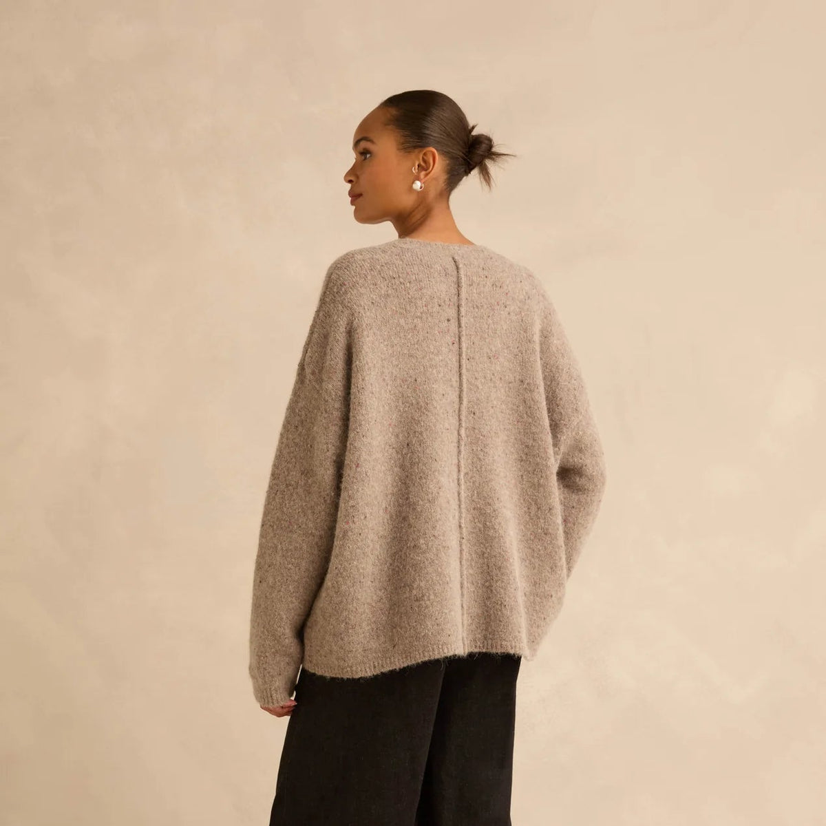 A model shown from the back wearing a light beige fuzzy knit sweater, black wide-leg trousers, and black boots, standing against a textured beige studio backdrop.