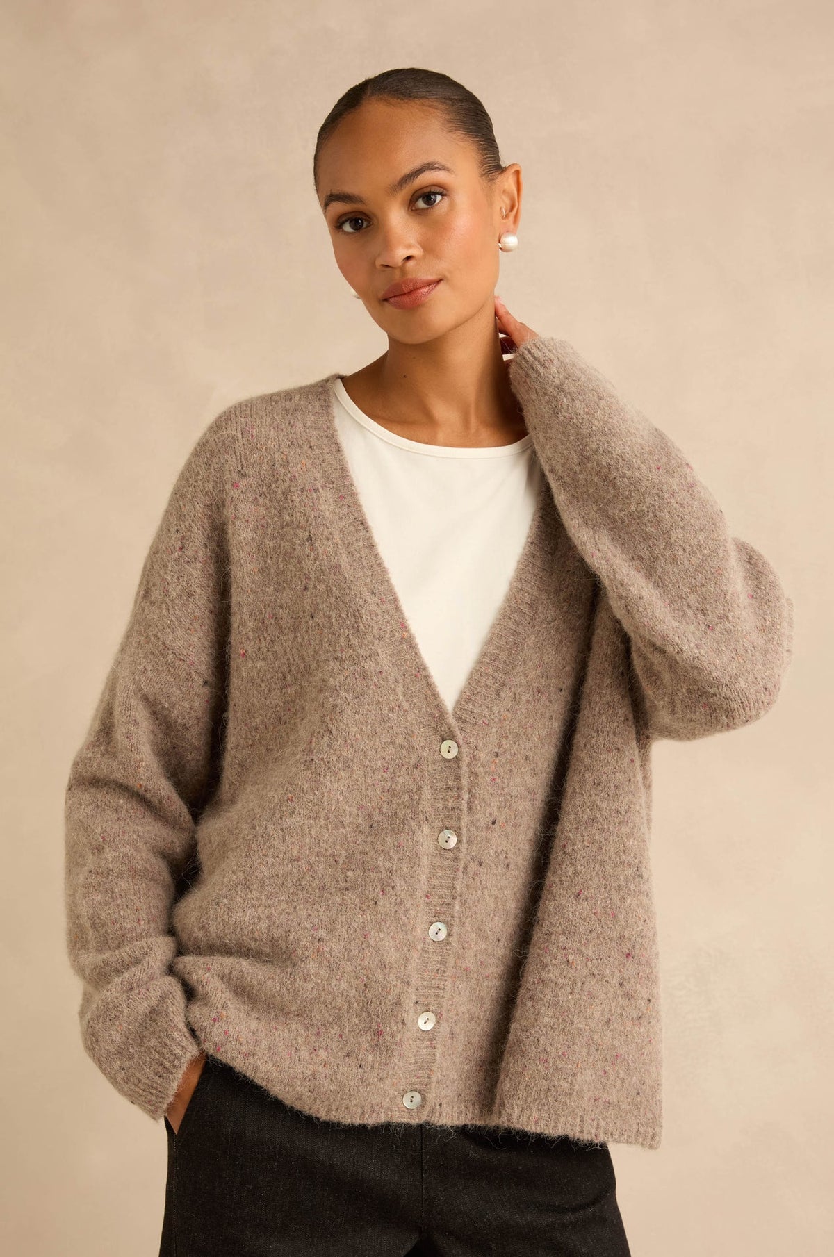 A woman wearing a fuzzy taupe knit cardigan over a white top and black pants, standing against a warm beige studio background.
