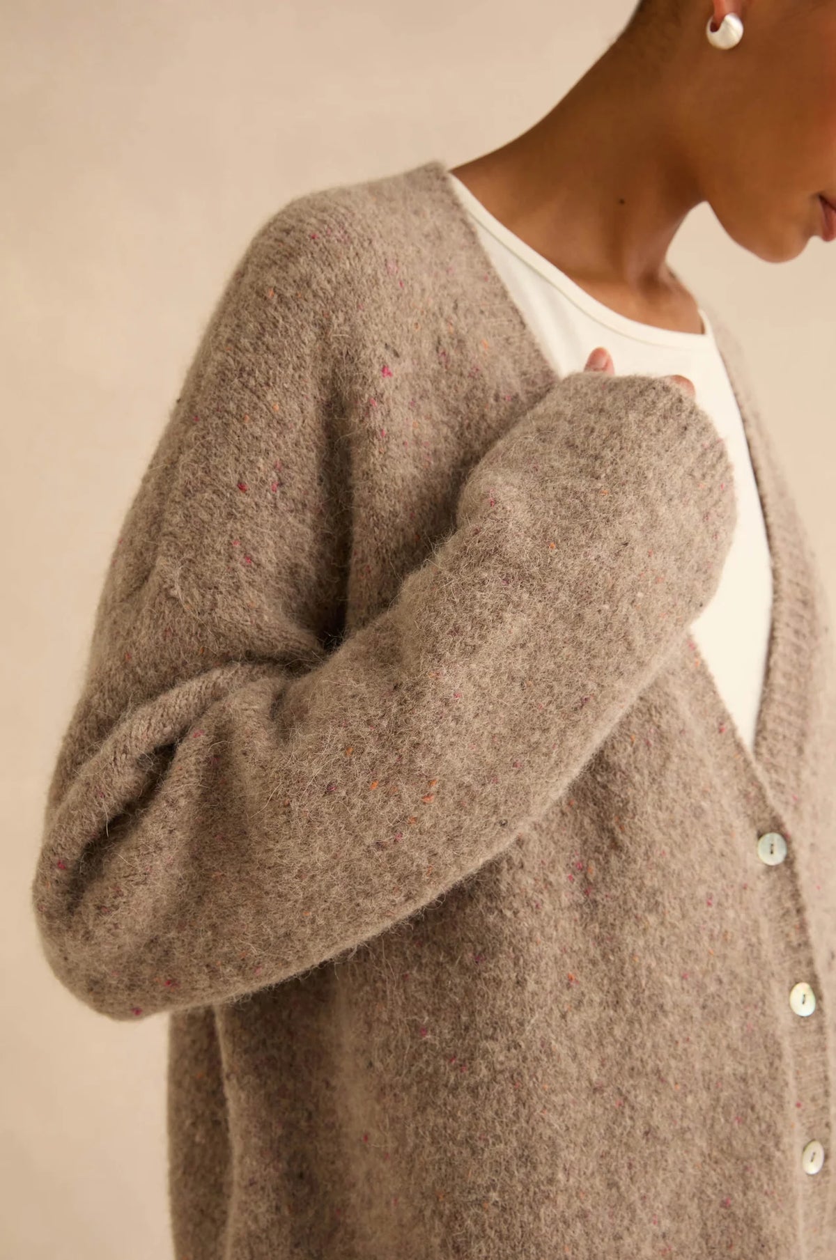 A close-up of a person wearing a fuzzy, oversized taupe-brown cardigan over a white top, against a plain beige background.