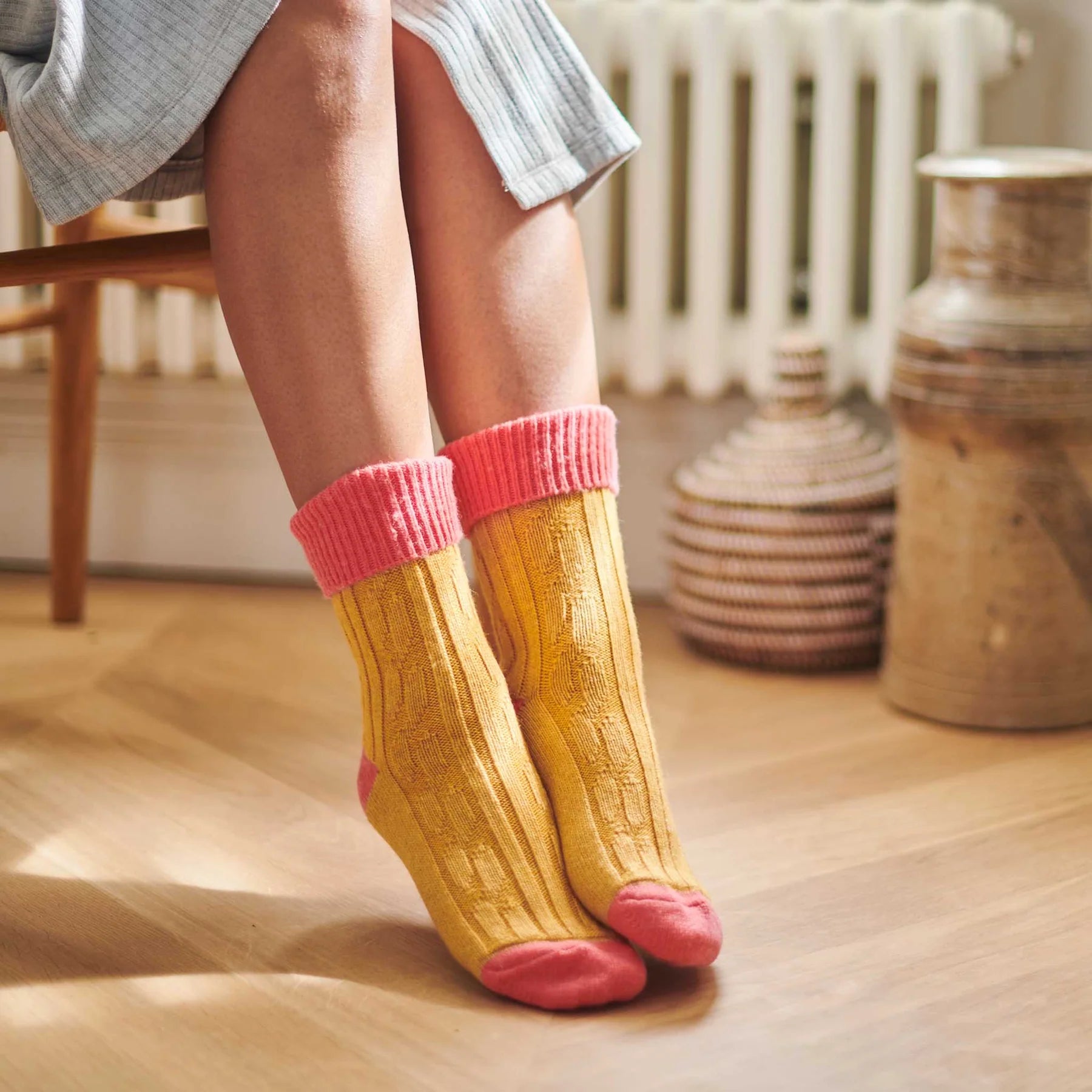 A person wearing yellow cable-knit socks with bright pink cuffs, heels, and toes, standing on a light wood floor in a cozy indoor setting.