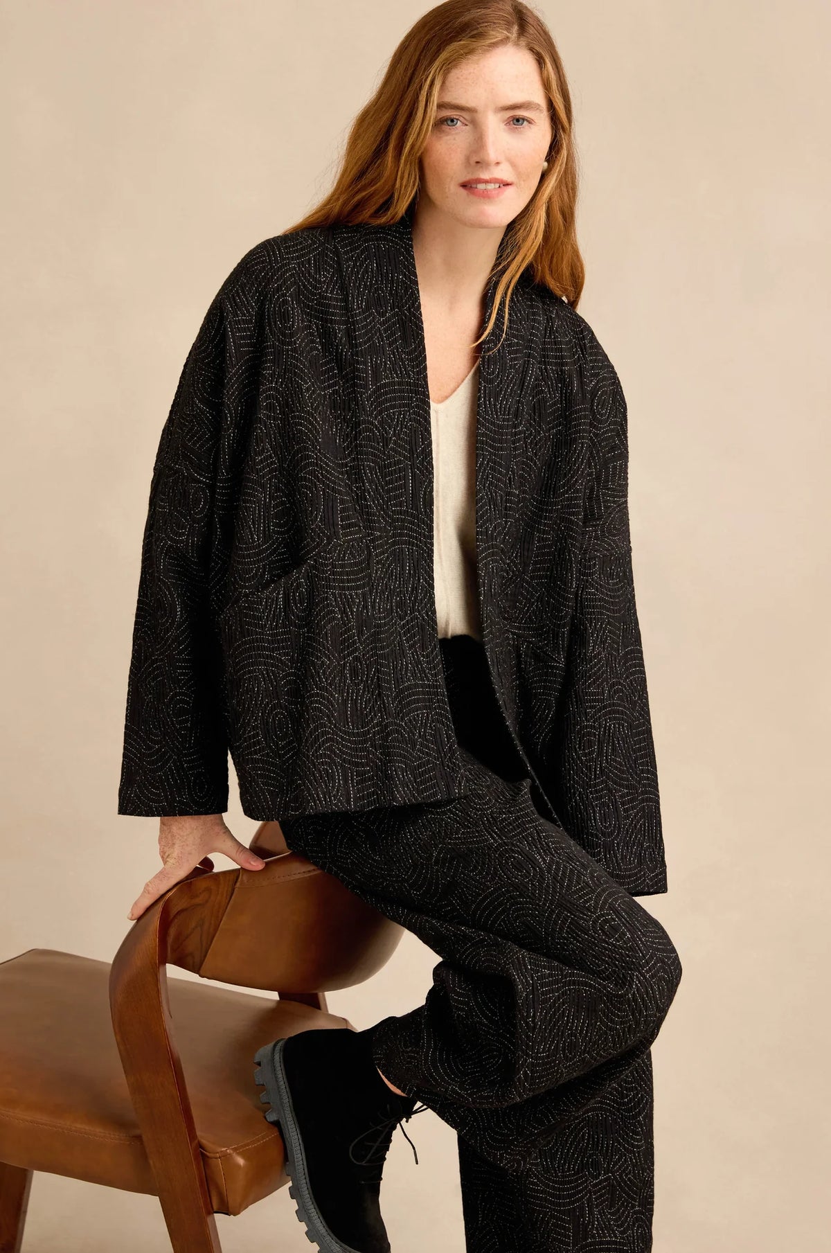 A woman models a matching black patterned jacket and trousers set, sitting on a wooden chair against a neutral beige background.