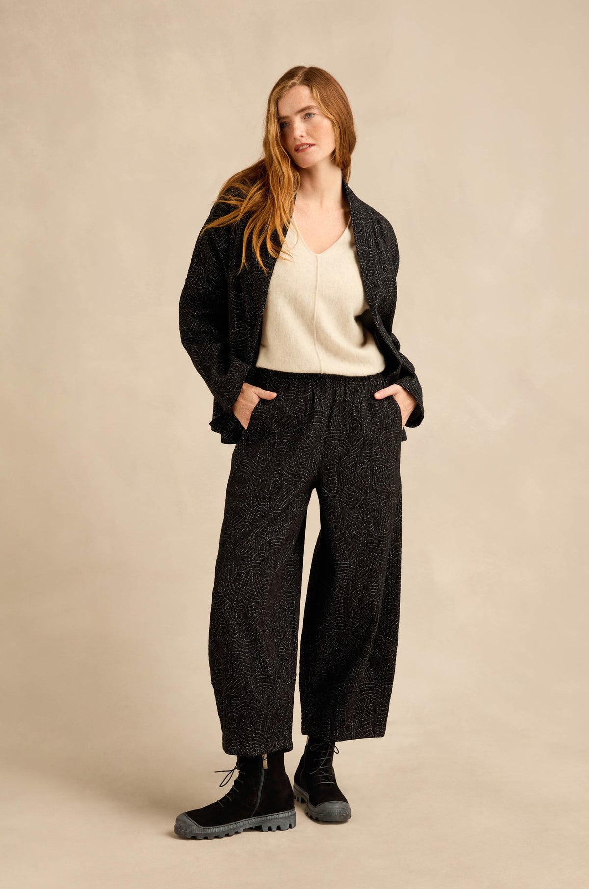 A woman stands against a warm beige studio backdrop wearing a dark textured cardigan, cream knit top, wide-leg dark trousers, and black lace-up boots.