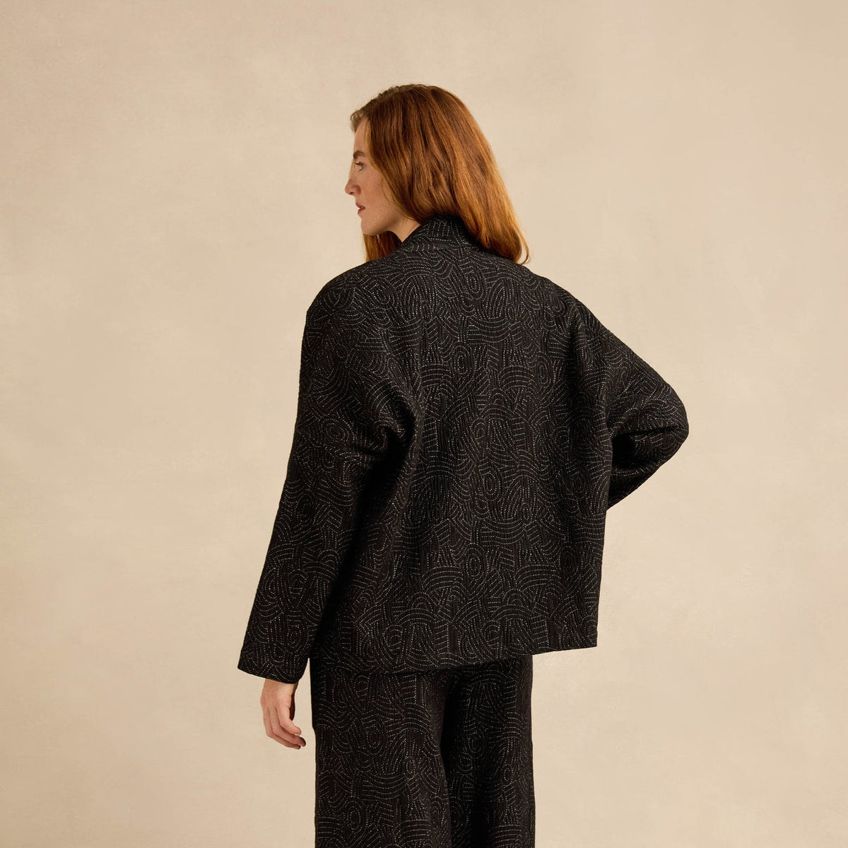 A model shown from the back wearing a matching dark charcoal gray textured jacket and wide-leg trousers set, standing against a solid beige studio background.
