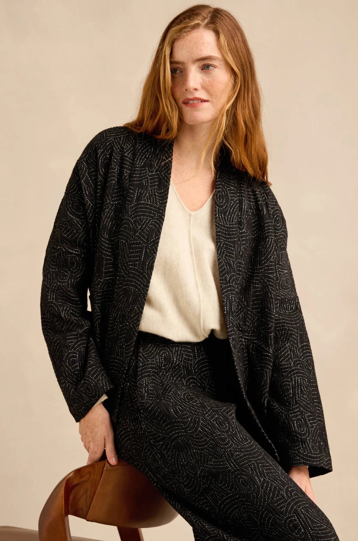 A woman models a matching black and gray patterned jacket and trousers set, worn over a cream-colored top, seated on a brown leather stool against a warm beige background.