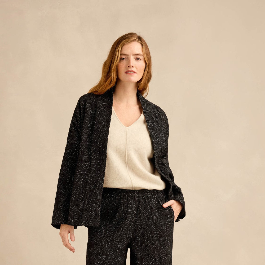 A woman models a dark charcoal gray textured knit set consisting of a cardigan and wide-leg trousers, styled with a cream V-neck knit top and black boots against a warm beige studio backdrop.