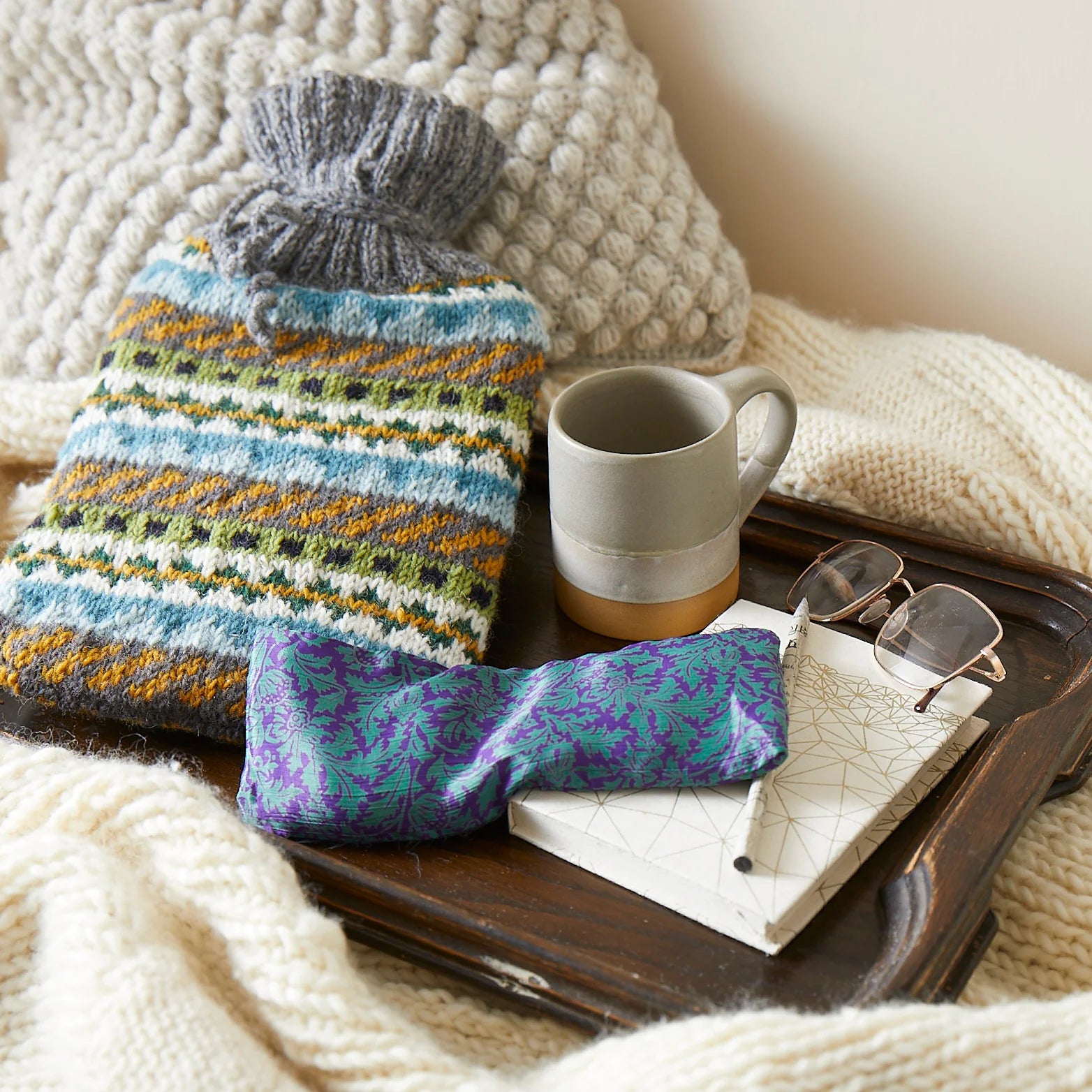 Fair isle patterned knitted hot water bottle, teal and purple eye mask, and a grey mug on a wooden tray.