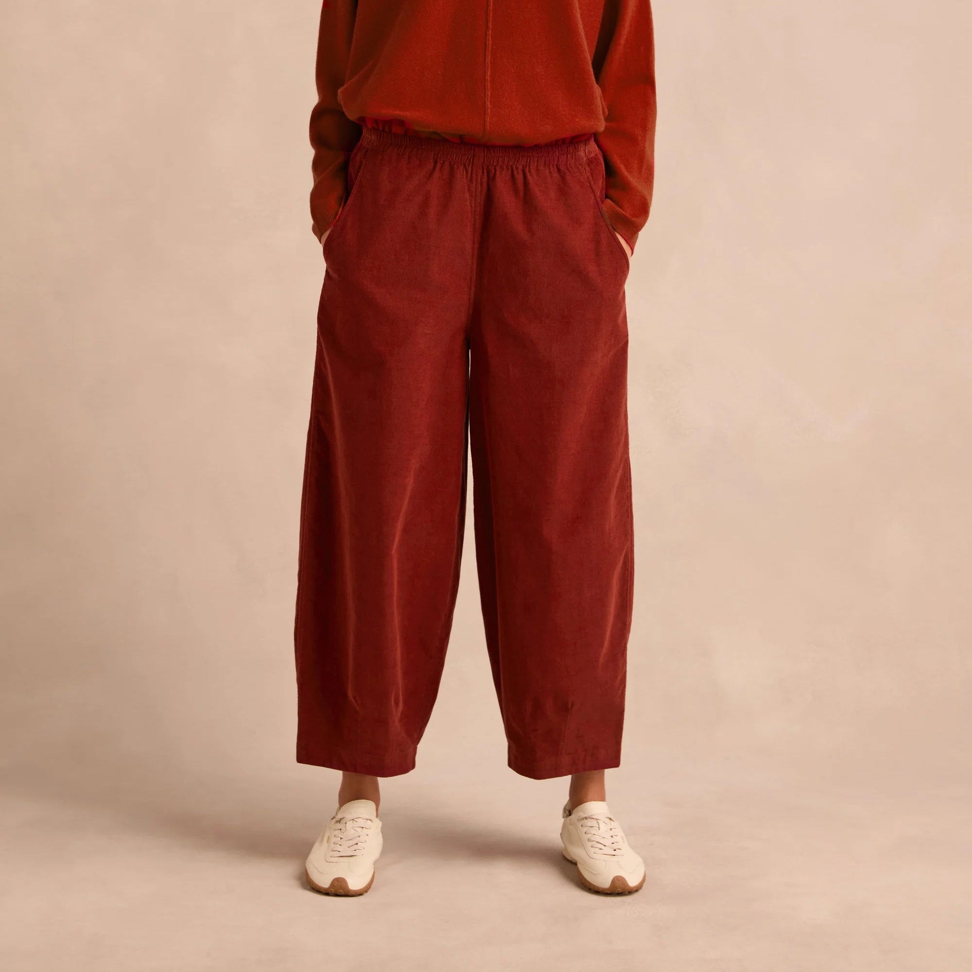 A woman modeling a rust-red cowl-neck knit sweater and matching wide-leg corduroy pants against a warm beige studio backdrop.