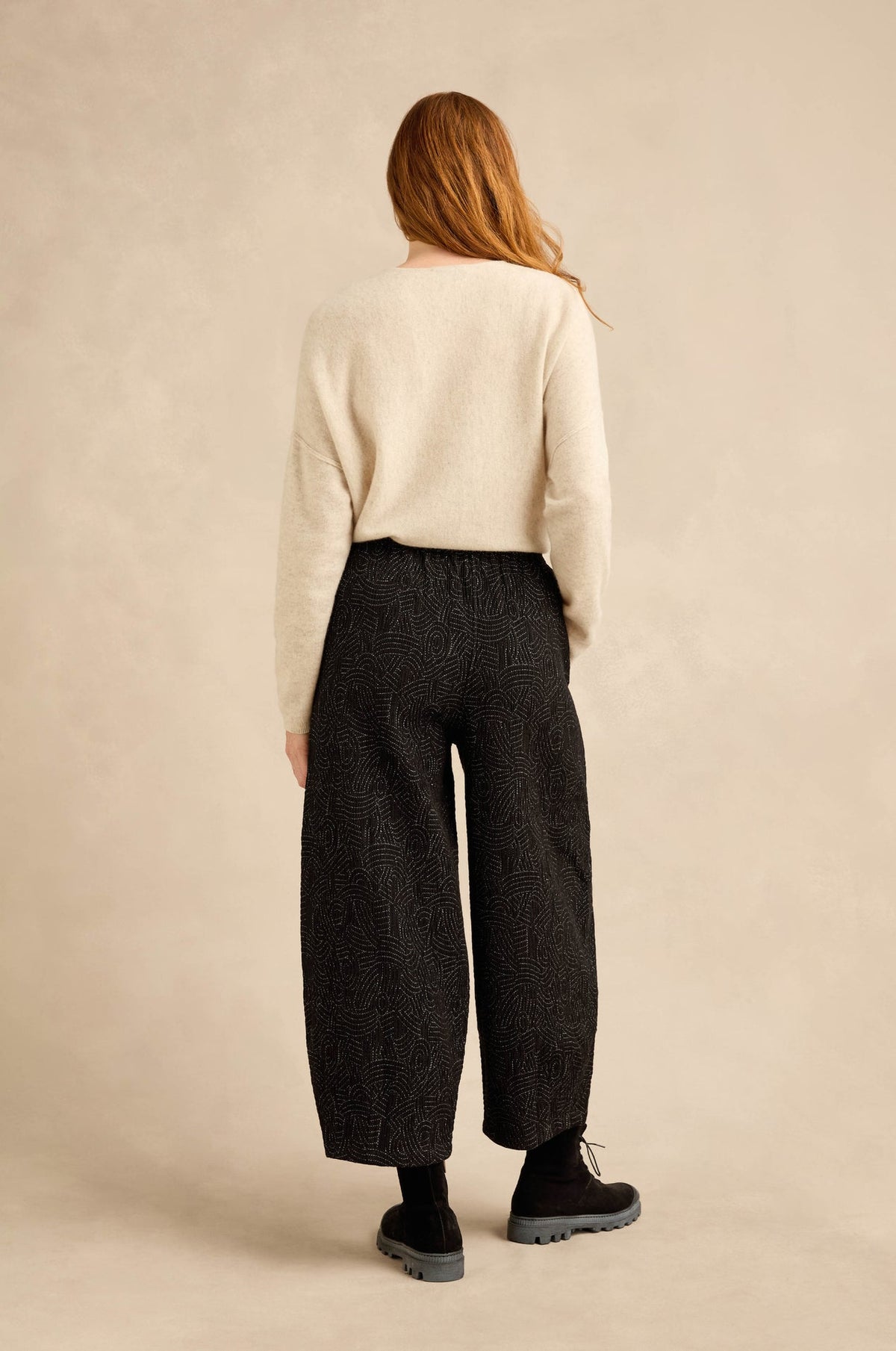 A woman is shown from behind wearing a cream knit sweater and dark charcoal wide-leg cropped trousers, standing against a neutral beige textured studio backdrop.