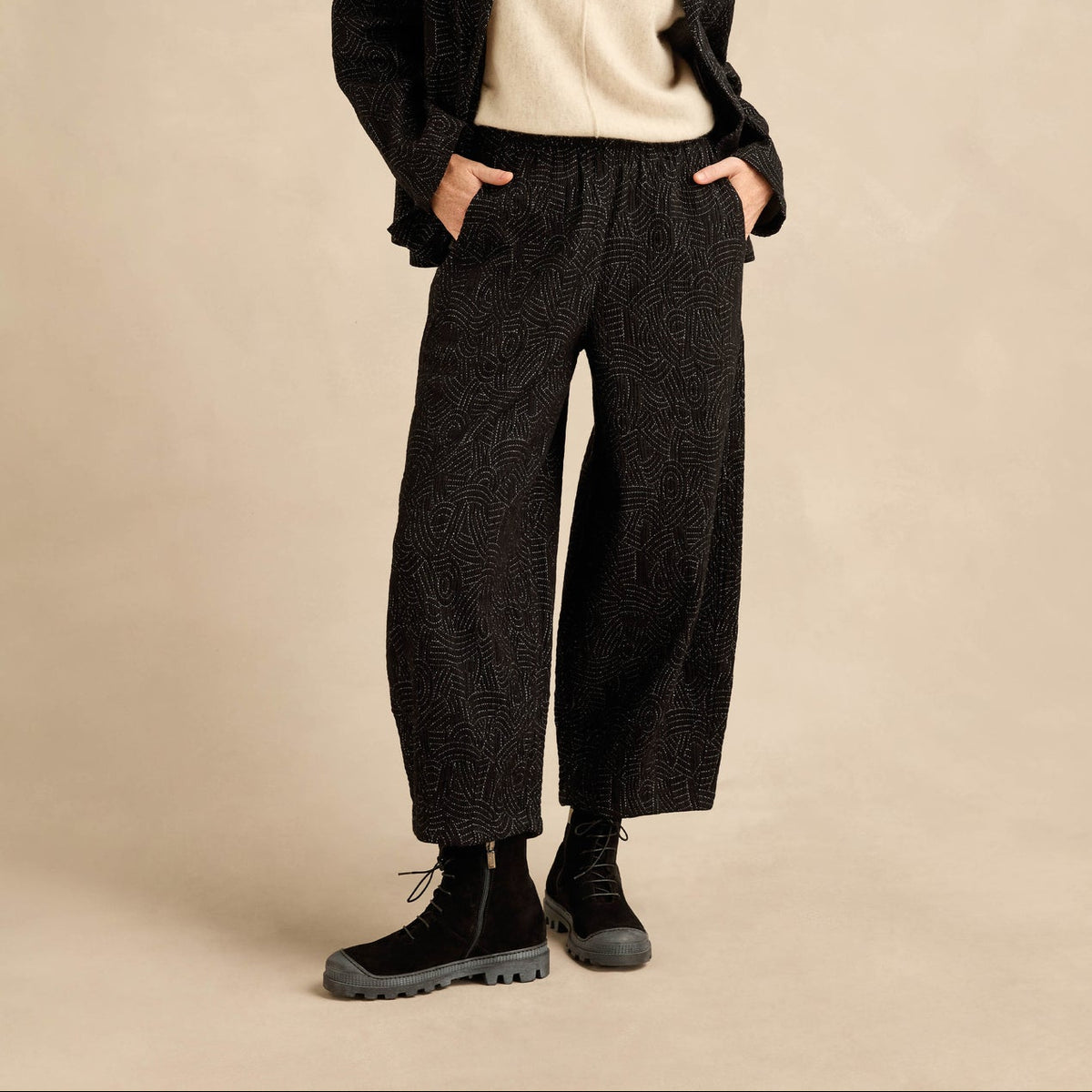 A woman stands against a warm beige studio backdrop wearing a dark textured cardigan, cream knit top, wide-leg dark trousers, and black lace-up boots.