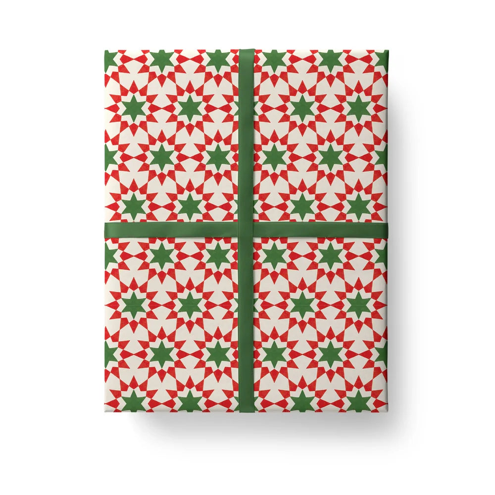 A rectangular gift box wrapped in festive holiday paper with a repeating red and green geometric star pattern on a white background, tied with a dark green ribbon in a cross shape, shown on a clean white background.