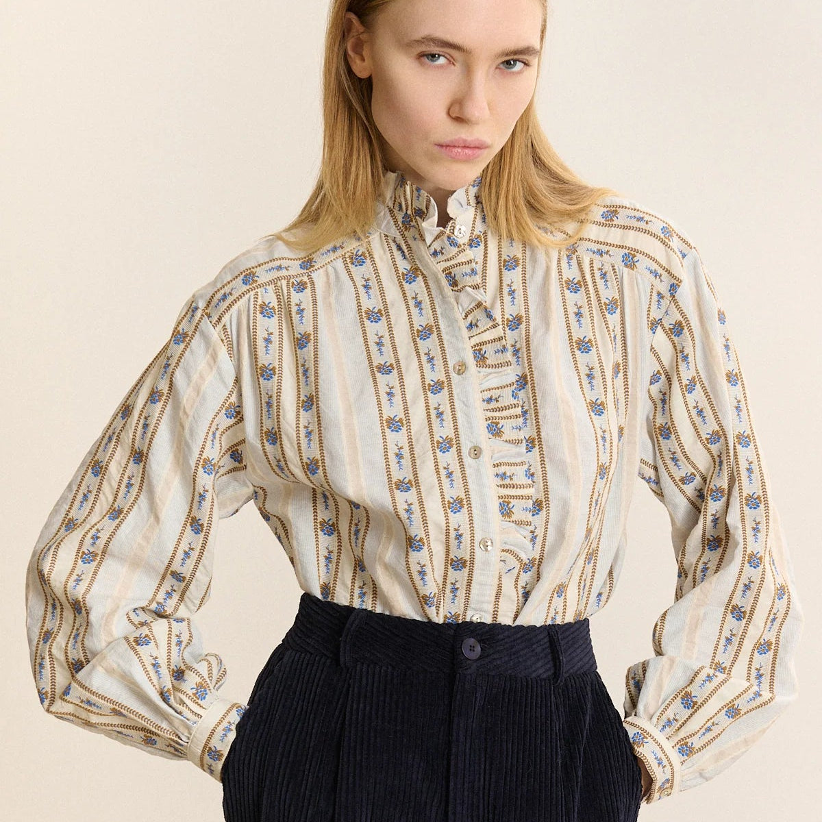 A woman modeling a cream-colored long-sleeve blouse with vertical blue and gold patterned stripes, paired with dark navy corduroy trousers, against a plain beige background.