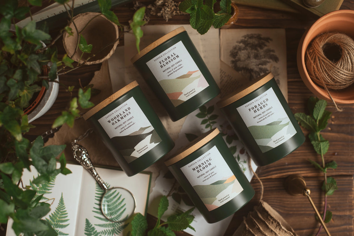 Four dark green candle jars with light wood lids are arranged on a rustic wooden surface with botanical props, including fresh green leaves, a magnifying glass, twine, and vintage-style botanical paper.