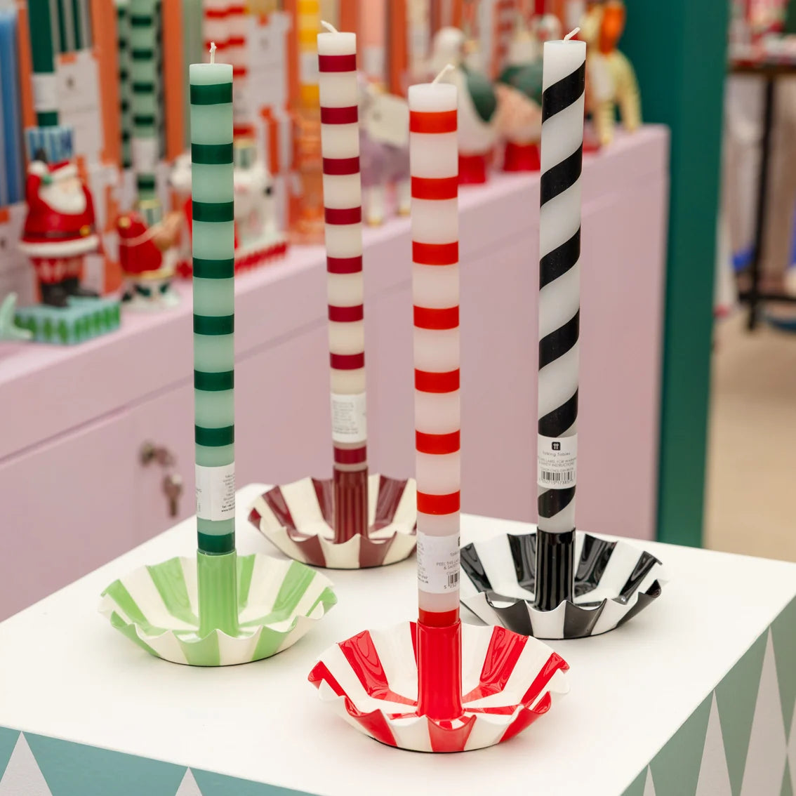 Four striped taper candles in paper cup holders, displayed on a white table in a store setting. The candles feature green and white, red and white, and black and white stripe patterns.