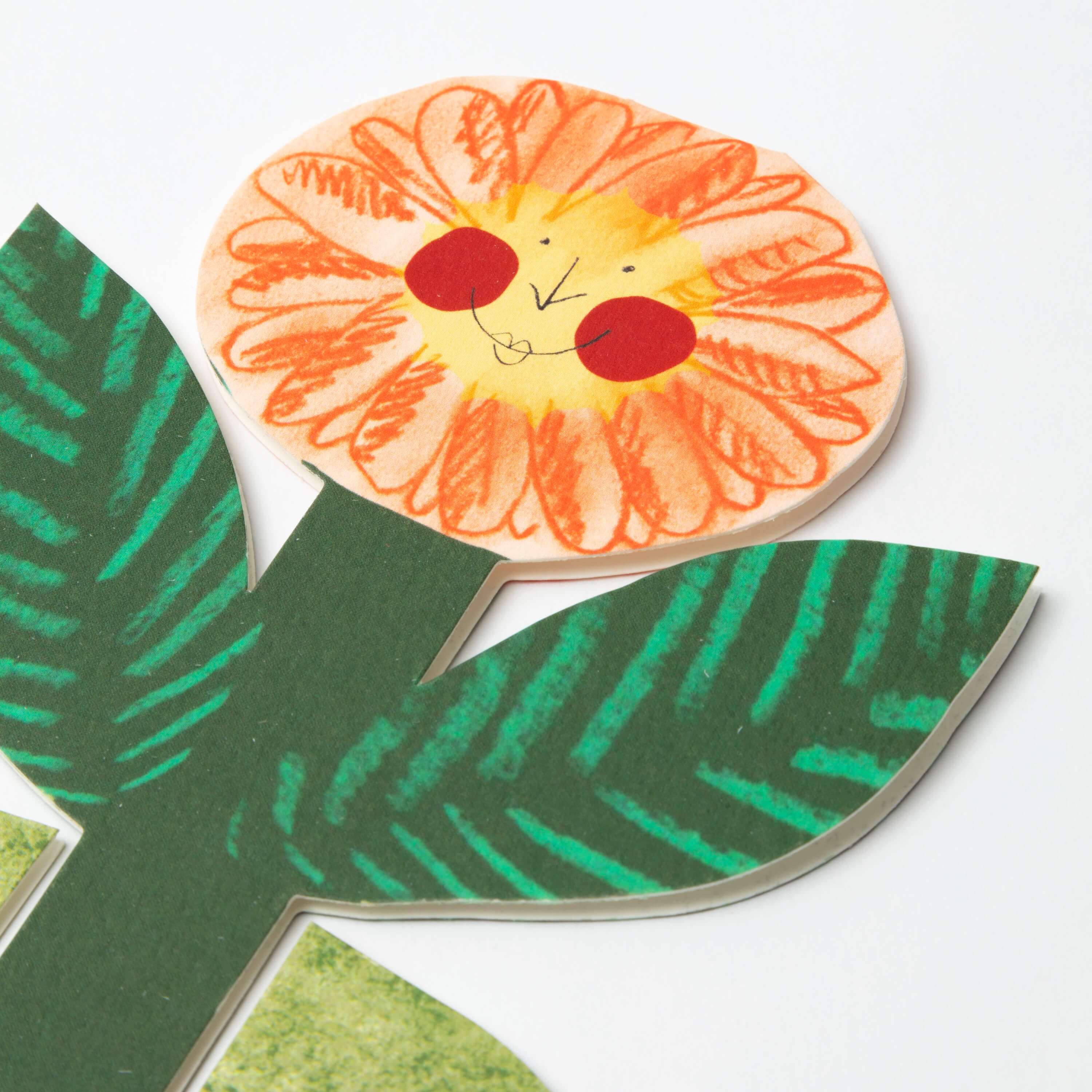 Close-up of a paper flower appliqué with an orange flower head featuring a smiling face and green leaves with teal stripes.