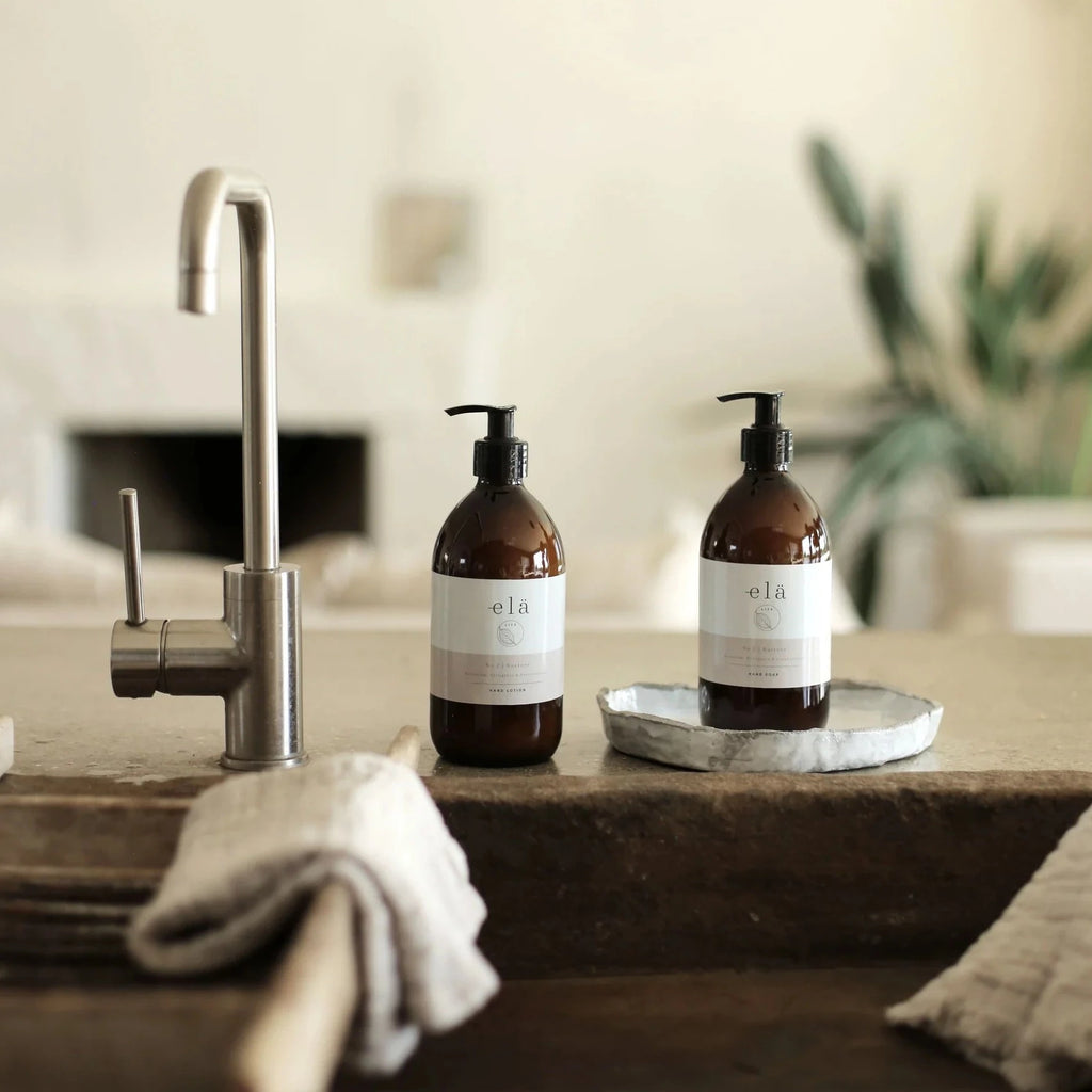two amber glass, pump dispensing bottles, containing No. 2 / NURTURE Hand & Body Lotion, by Elä Life, positioned next to a chrome mixer tap