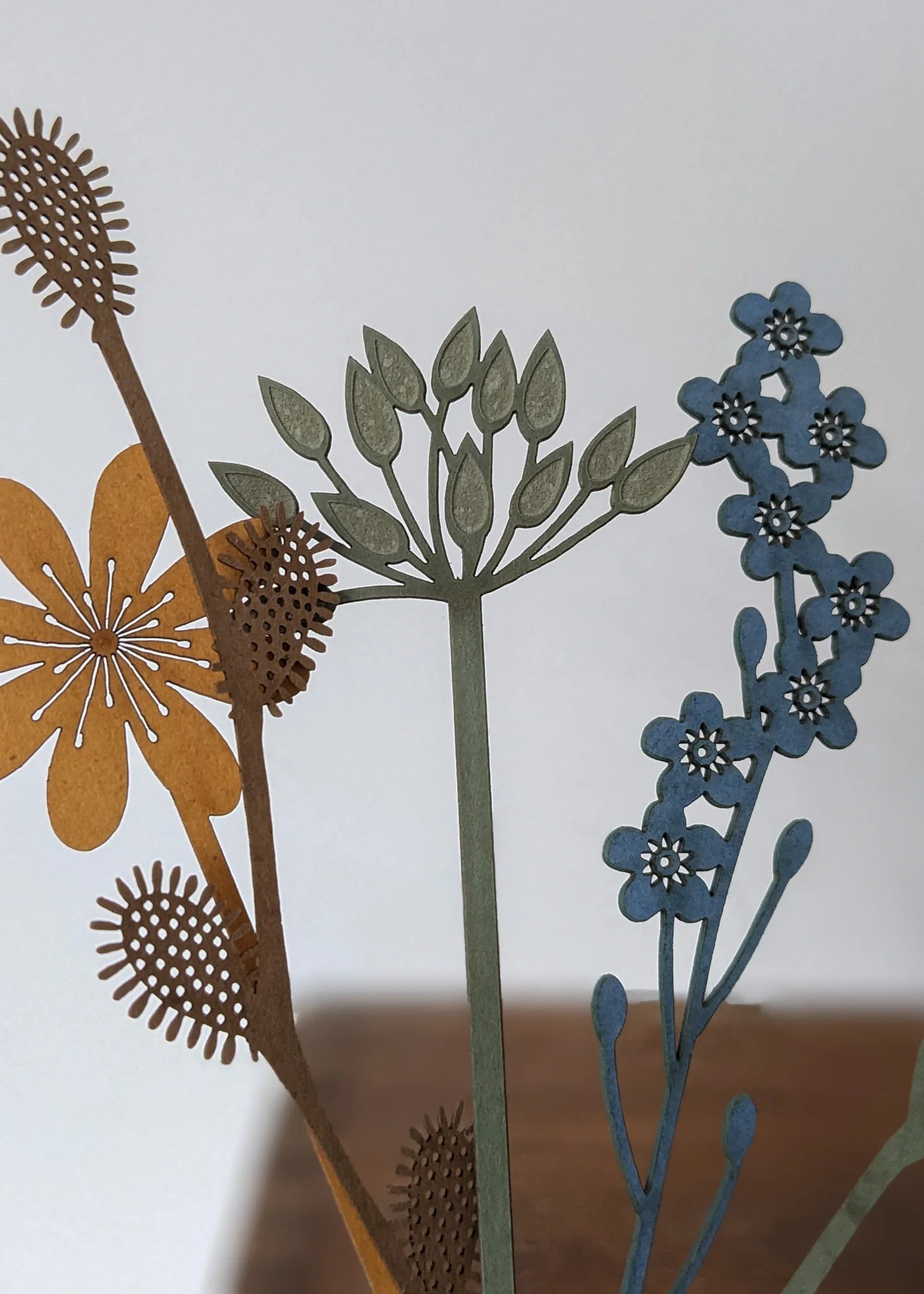 Intricate laser-cut floral arrangement featuring stylized flowers and stems in shades of brown, green, and blue.